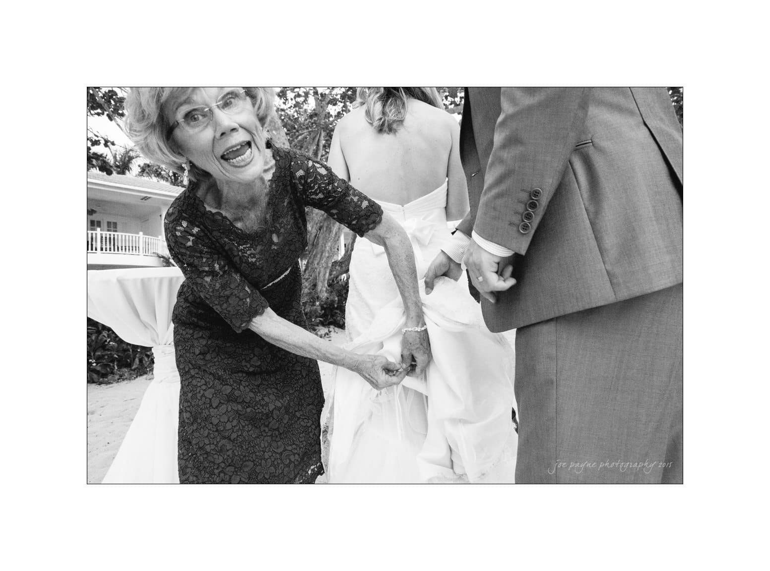 An elderly woman playfully interacts with a bride by adjusting her wedding dress, while a groom stands beside them. The scene is lively and candid, set outdoors near a building.