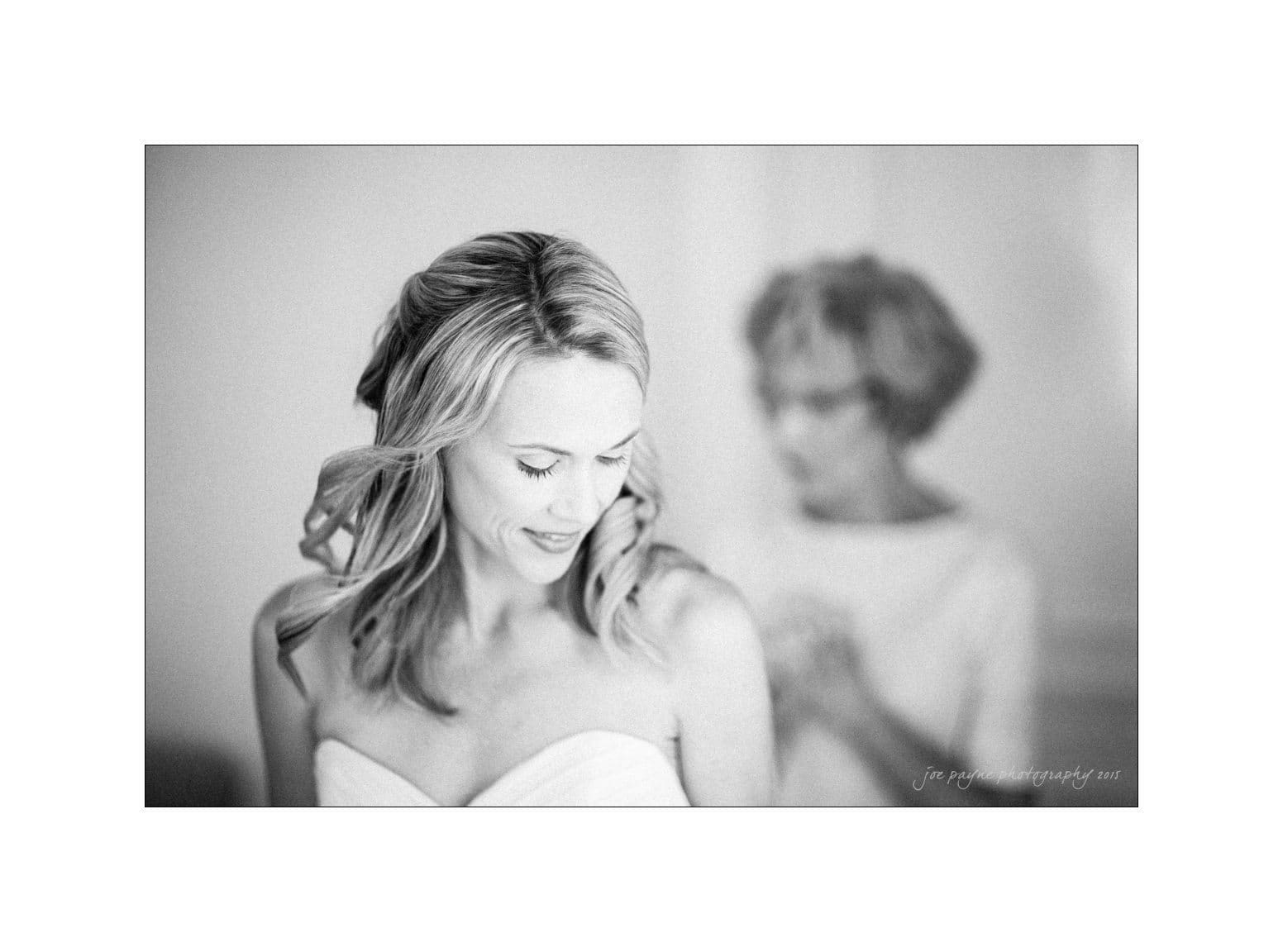 A black-and-white photo of a smiling woman in a strapless top looking down. Her hair is styled in loose waves. In the blurred background, an older woman is visible, standing with her hands close together.