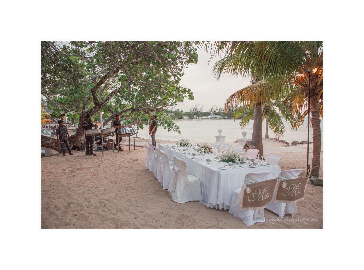 A beautifully set table for a wedding on a sandy beach, adorned with white tablecloths and floral centerpieces. Two chairs at the end have decorative initials. Musicians stand nearby, and palm trees frame the serene view of the water.