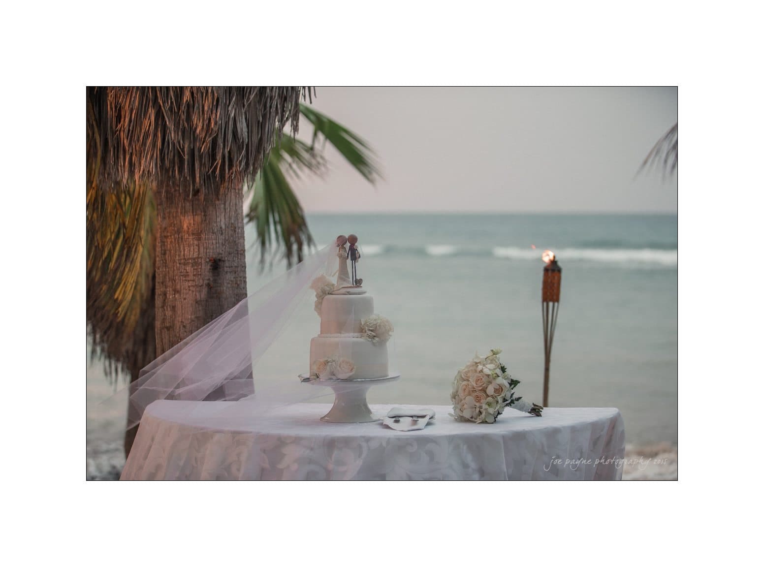 A three-tier white wedding cake with a bride and groom topper sits on a covered table by the beach. A bouquet and decorative floral arrangements are nearby, with palm leaves and the ocean in the background.