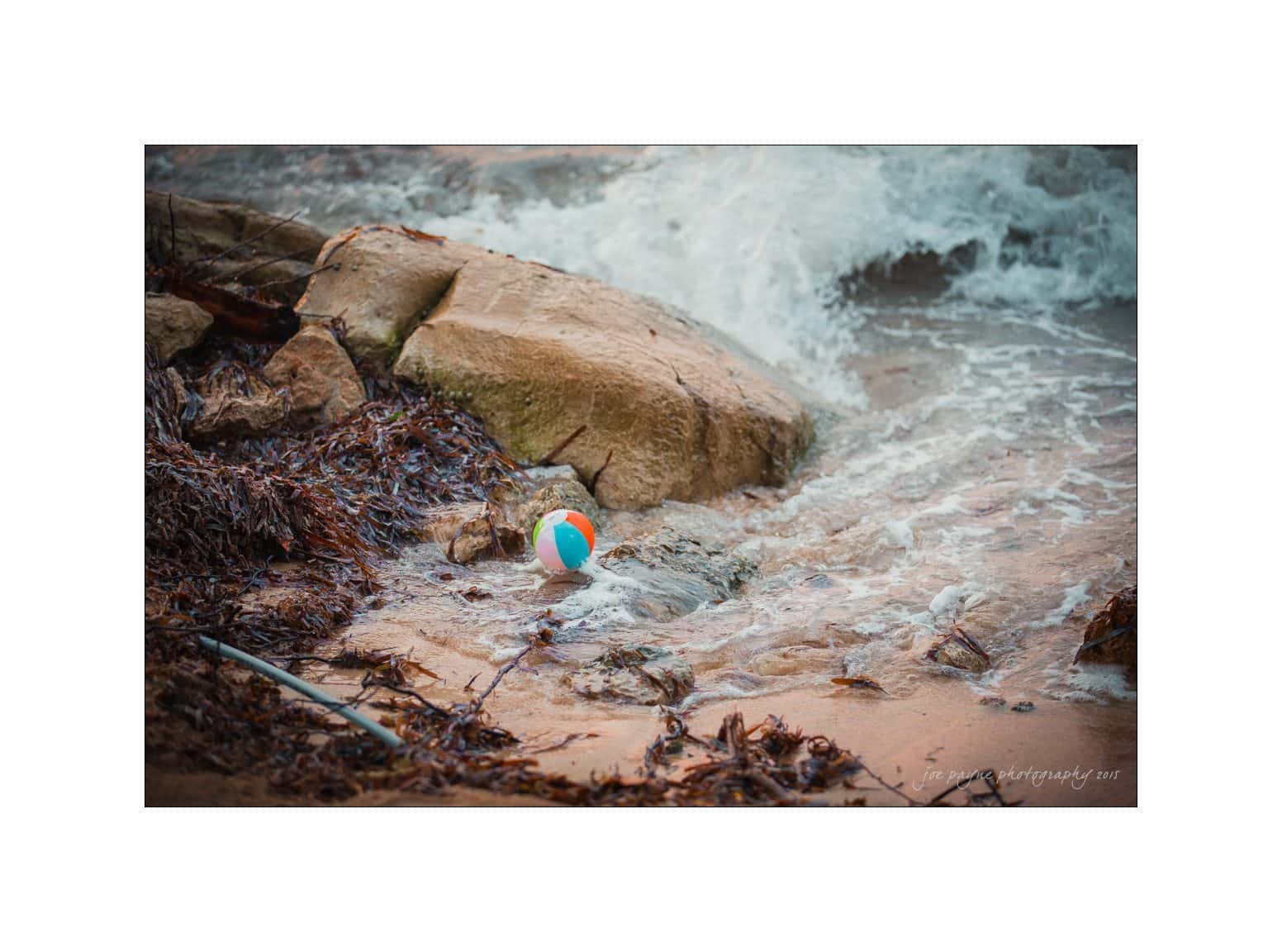 half moon montego bay wedding photography - leah & evan A colorful beach ball rests on a rocky shore, surrounded by seaweed and gentle waves. The surf is washing over the rocks, creating a dynamic and serene coastal scene.