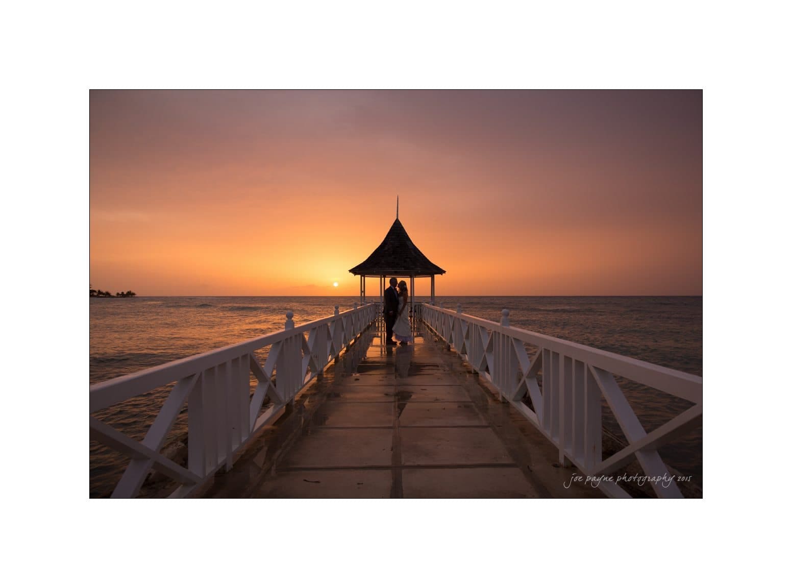 A person stands at the end of a wooden pier with a gazebo, silhouetted against a vibrant orange sunset over the ocean. The pier has white railings, and the sky is a gradient of warm colors.