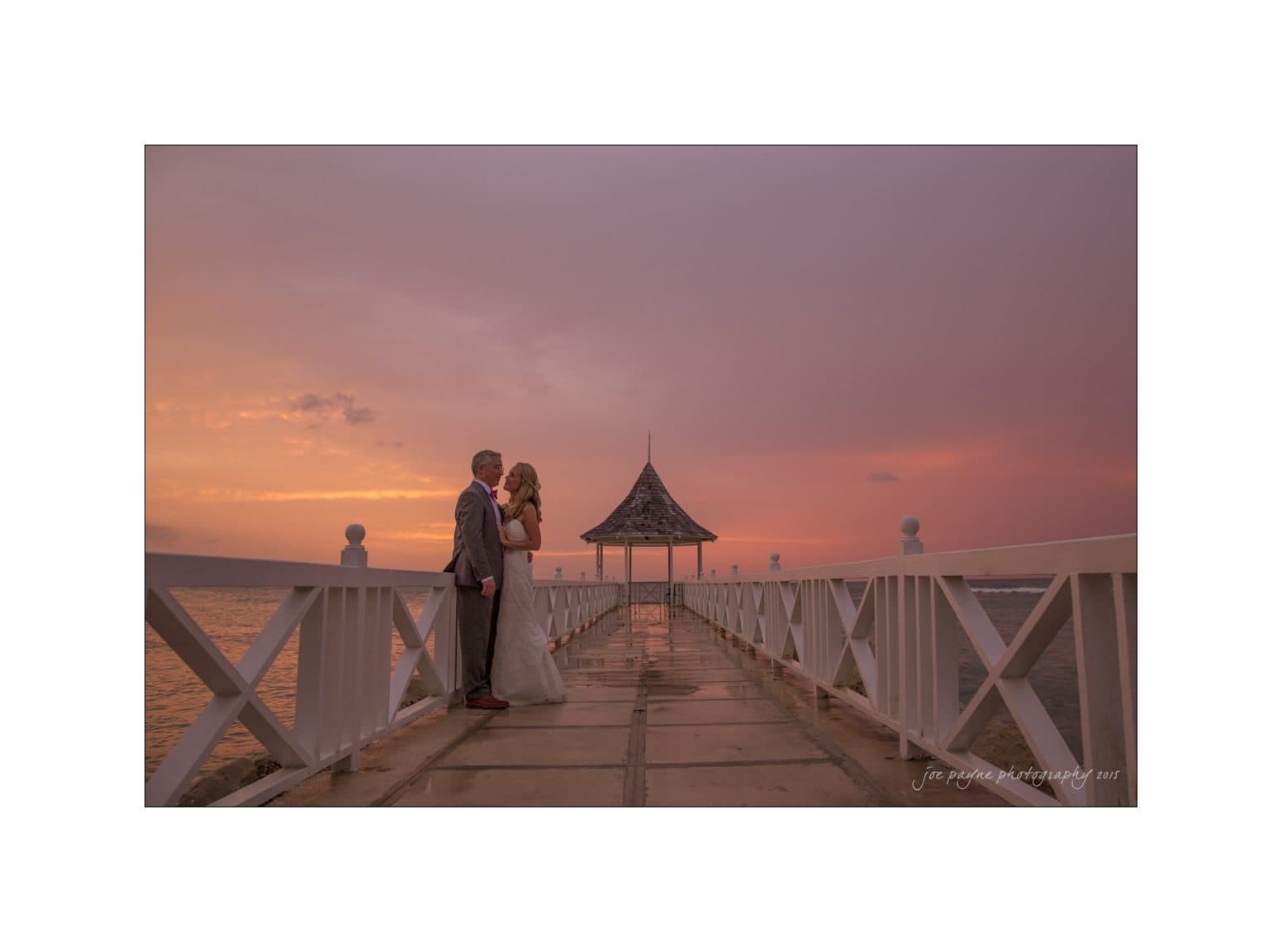 A couple embraces lovingly on a wooden pier at sunset. The sky is filled with warm hues of pink and orange, reflecting on the water. A gazebo is visible at the end of the pier.