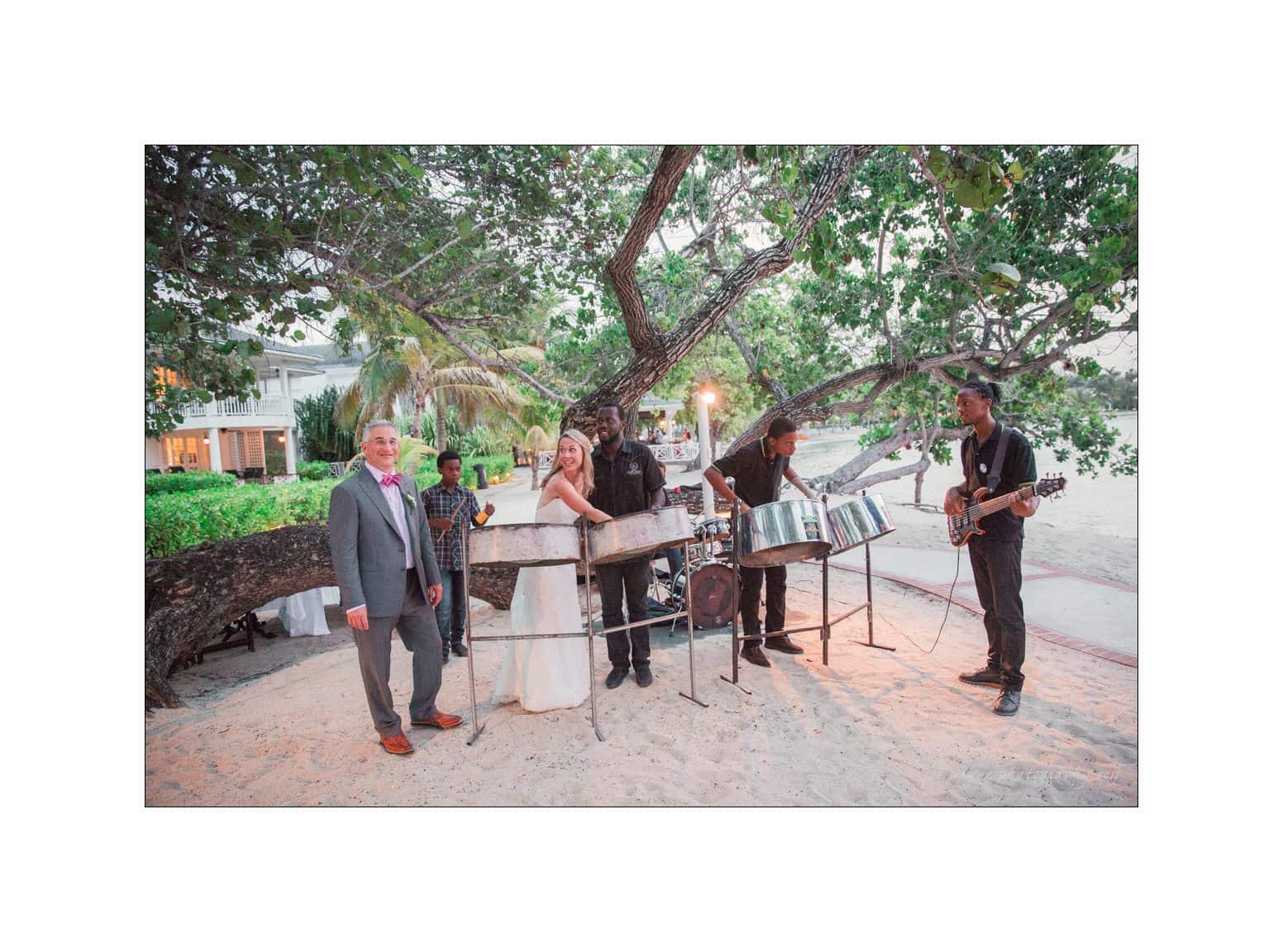 A bride and groom stand on a sandy beach beside a group of musicians playing steel drums and guitar under a large tree. The setting is tropical, with lush greenery and soft lighting in the background.