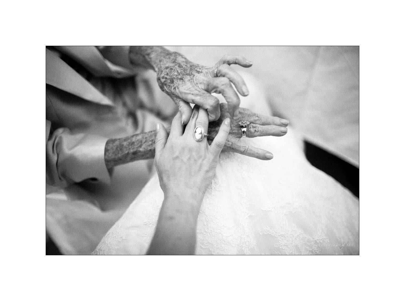 A black and white close-up photo of an elderly person and a younger person holding hands. The older person is placing a ring on the younger persons finger. They are wearing a suit and a lace garment, suggesting a formal occasion.