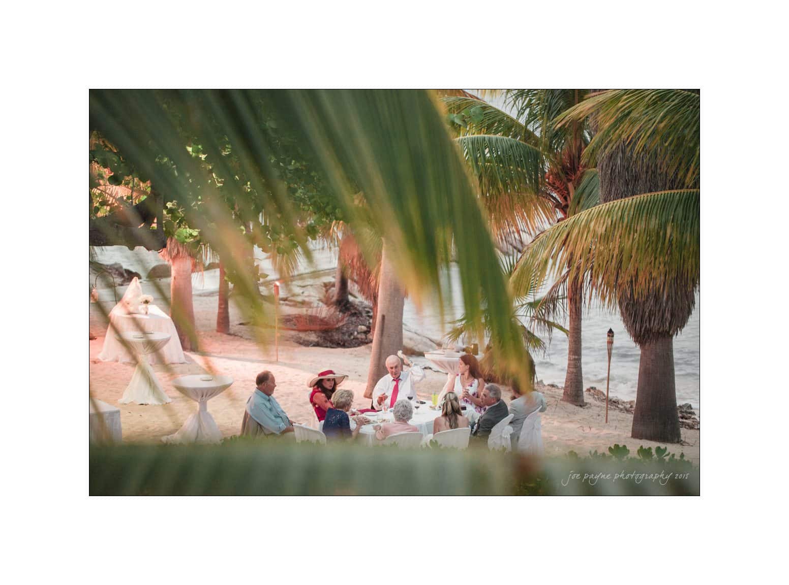 A group of people gathers around a table on a sandy beach surrounded by palm trees. The scene appears relaxed and tropical, with the ocean in the background.
