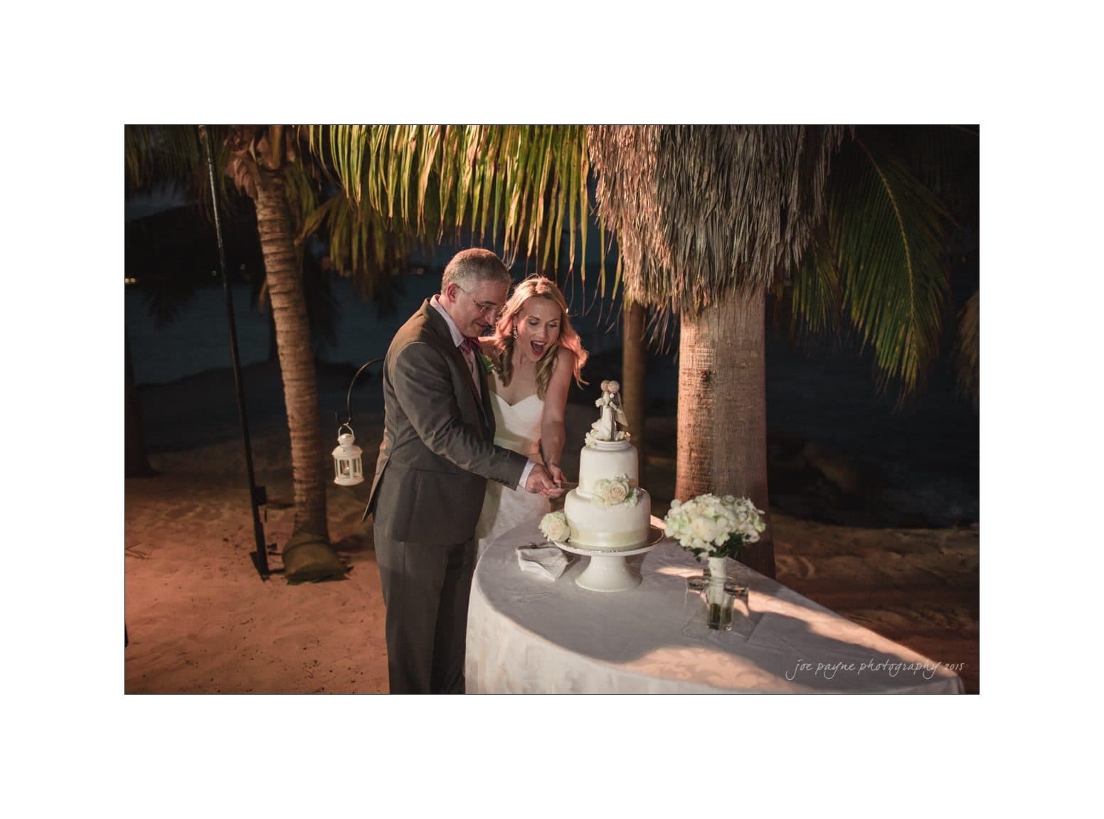 A bride and groom cut their wedding cake together on a beach at night. They are surrounded by palm trees and soft lighting, creating a romantic atmosphere. A bouquet of white flowers is on the table.