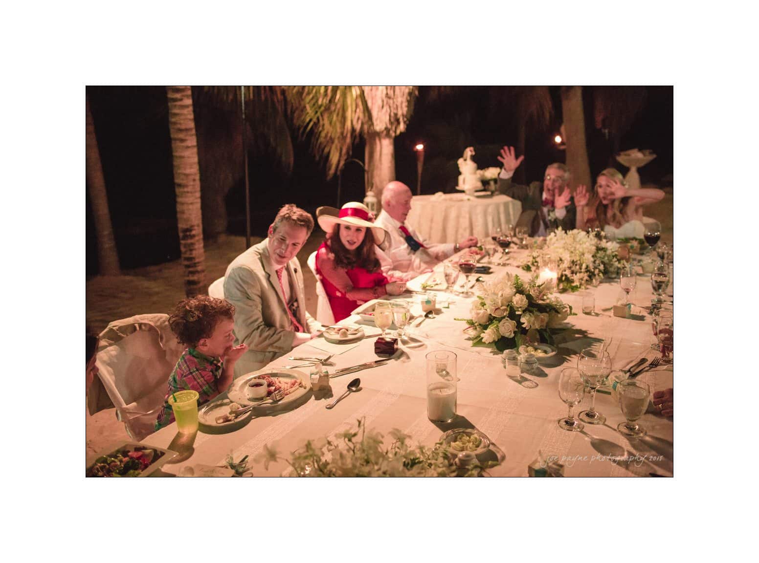 A group of people sit around an elegantly decorated dinner table outdoors at night. The table is adorned with white flowers and candles. Some guests are engaged in conversation, while others smile and look toward the camera.