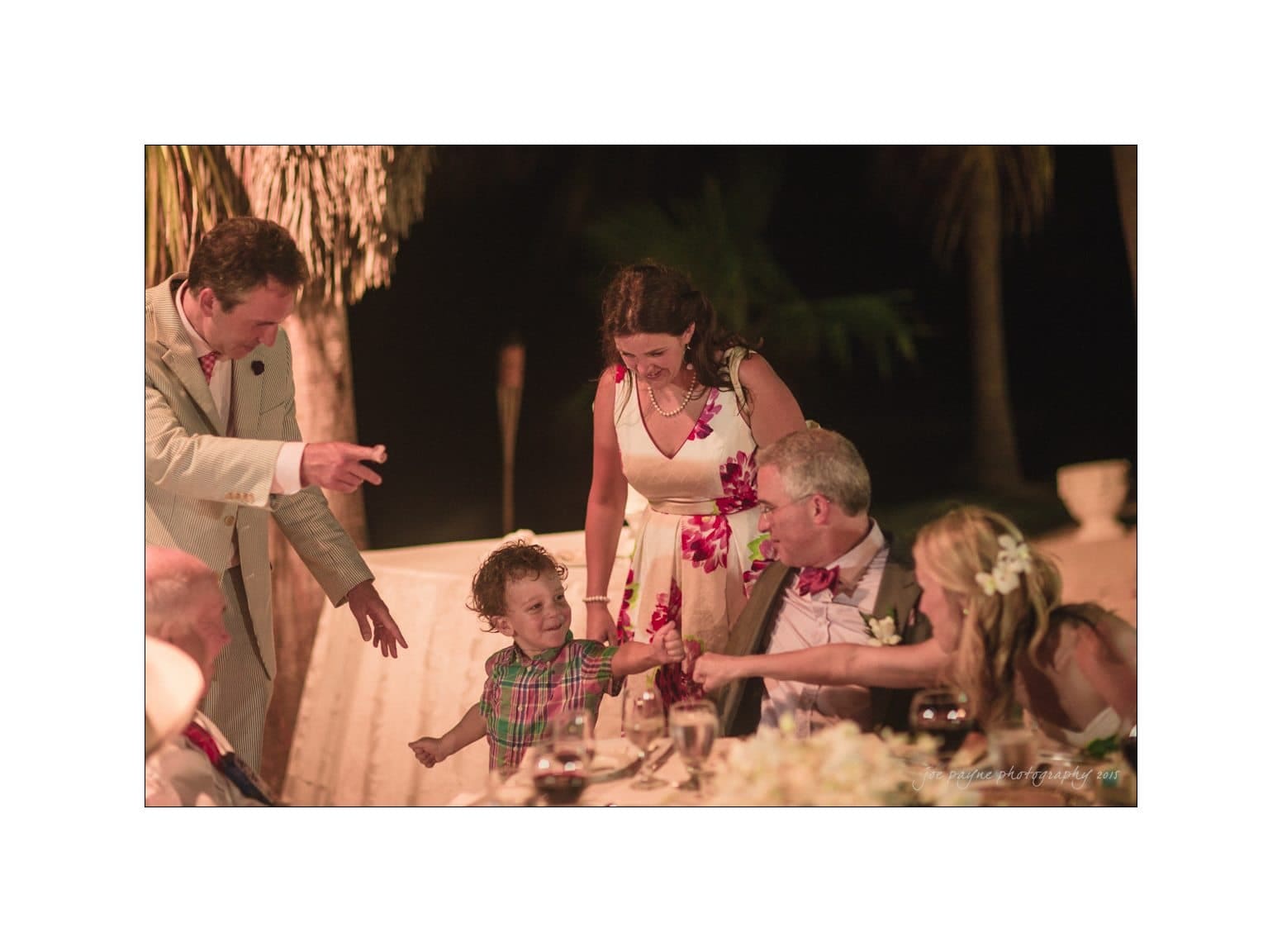 A group of people enjoying an outdoor evening event. A young child smiles and reaches for an older mans hand. Two women and a man stand nearby, looking on with joy. The setting is warmly lit, with palm tree shadows in the background.