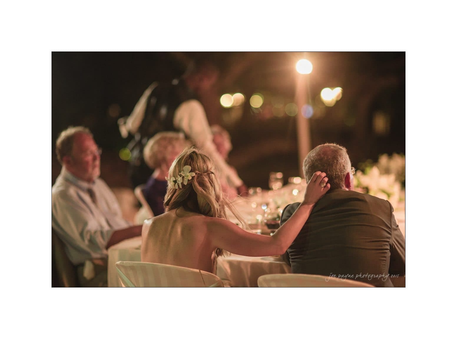 A woman with a flower in her hair affectionately touches the shoulder of an older man at an outdoor nighttime event. They sit at a table with other attendees, under soft lighting with blurred lights in the background.