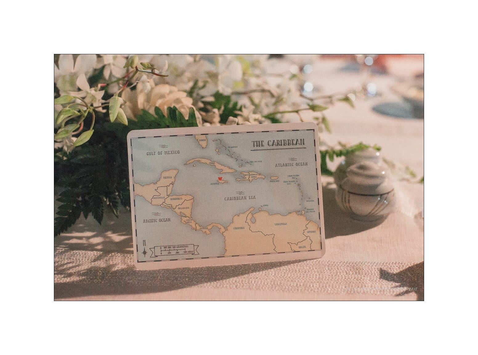 A decorative card on a table displaying a map of the Caribbean, surrounded by white flowers and soft lighting. The card identifies bodies of water like the Gulf of Mexico and the Atlantic Ocean. Tableware is partially visible in the background.