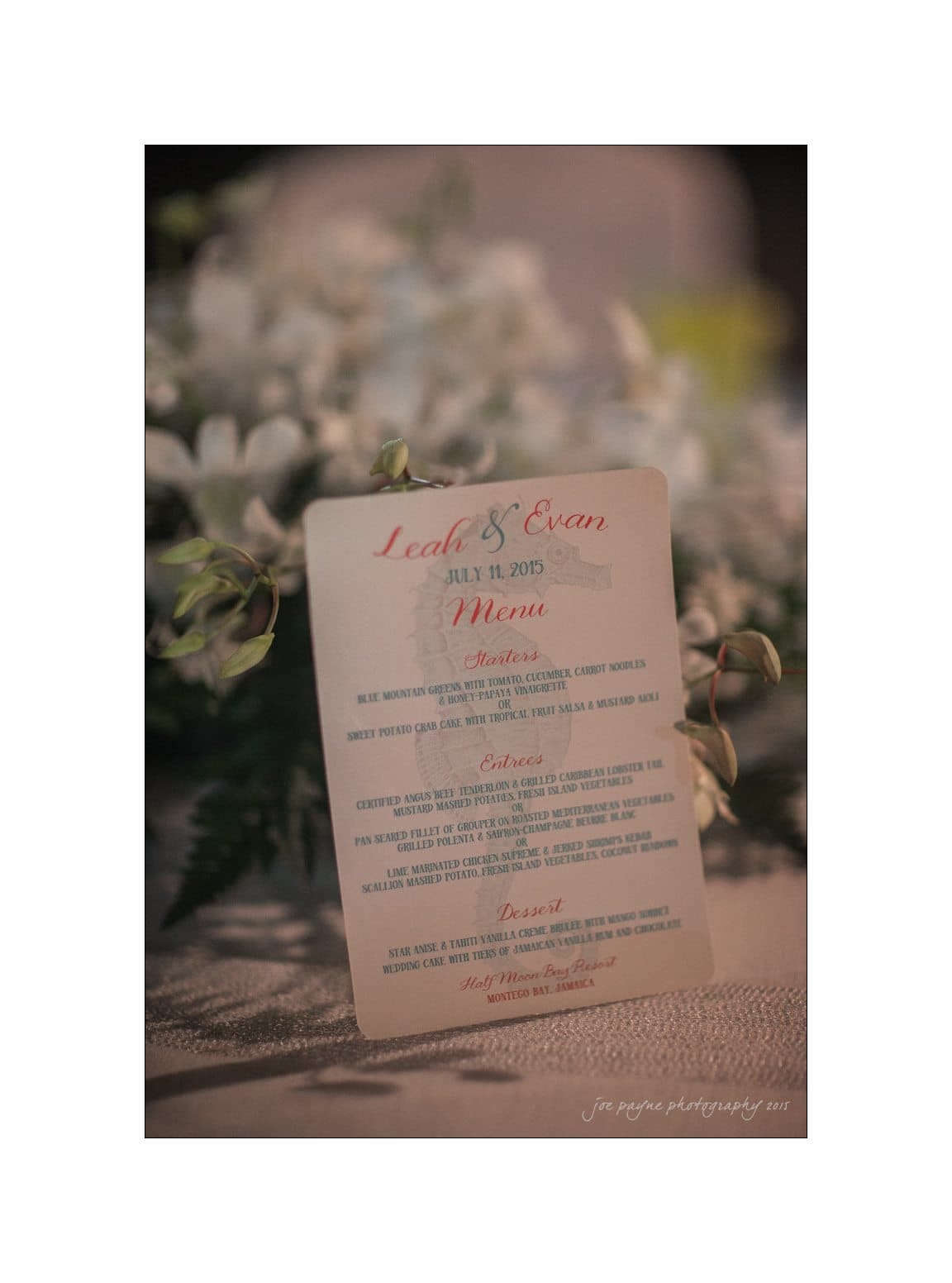 A wedding menu card for Leah and Evan, dated July 11, 2015, placed on a table with white flowers in the background.