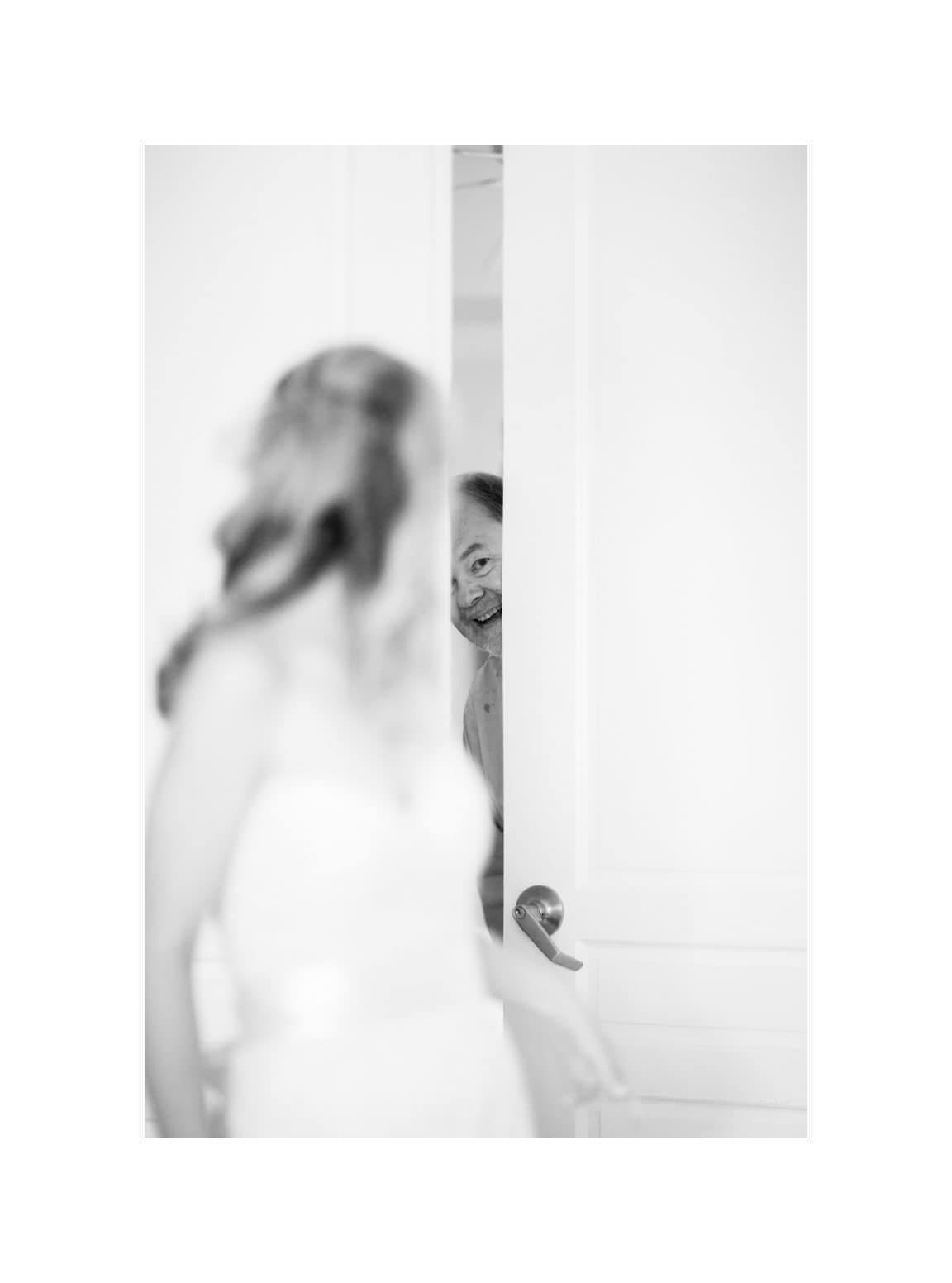 A black and white photo shows a bride in a wedding dress facing a slightly open door. A smiling man peeks through the doorway. The focus is on his joyful expression.