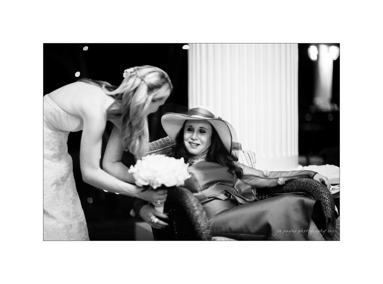 A woman in a wedding dress leans toward a seated woman in a wide-brimmed hat, offering a bouquet. The seated woman smiles warmly. The image is in black and white with a soft focus on the subjects.