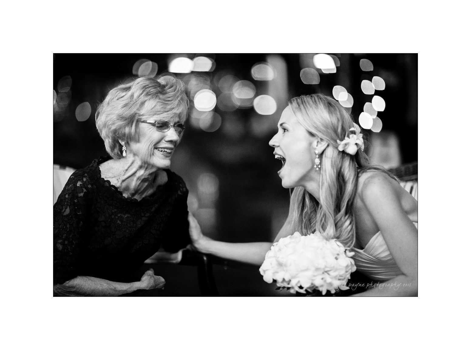 A black and white photo of a bride laughing joyfully with an elderly woman. The bride, holding a bouquet, is seated next to the woman, both sharing a moment of happiness against a blurred, twinkling background.