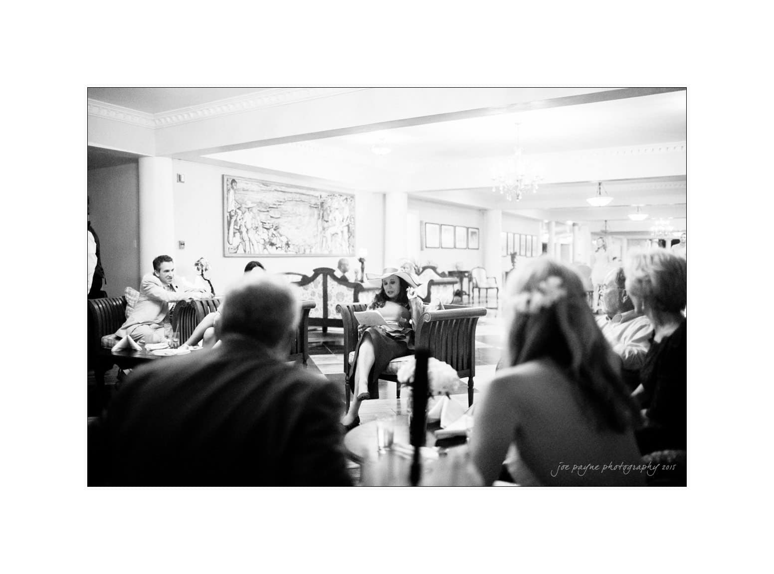A black and white photo capturing a group of people seated in a lounge area. A woman in the center holds a microphone, possibly speaking or singing. The setting is relaxed, with several attendees engaged and listening.