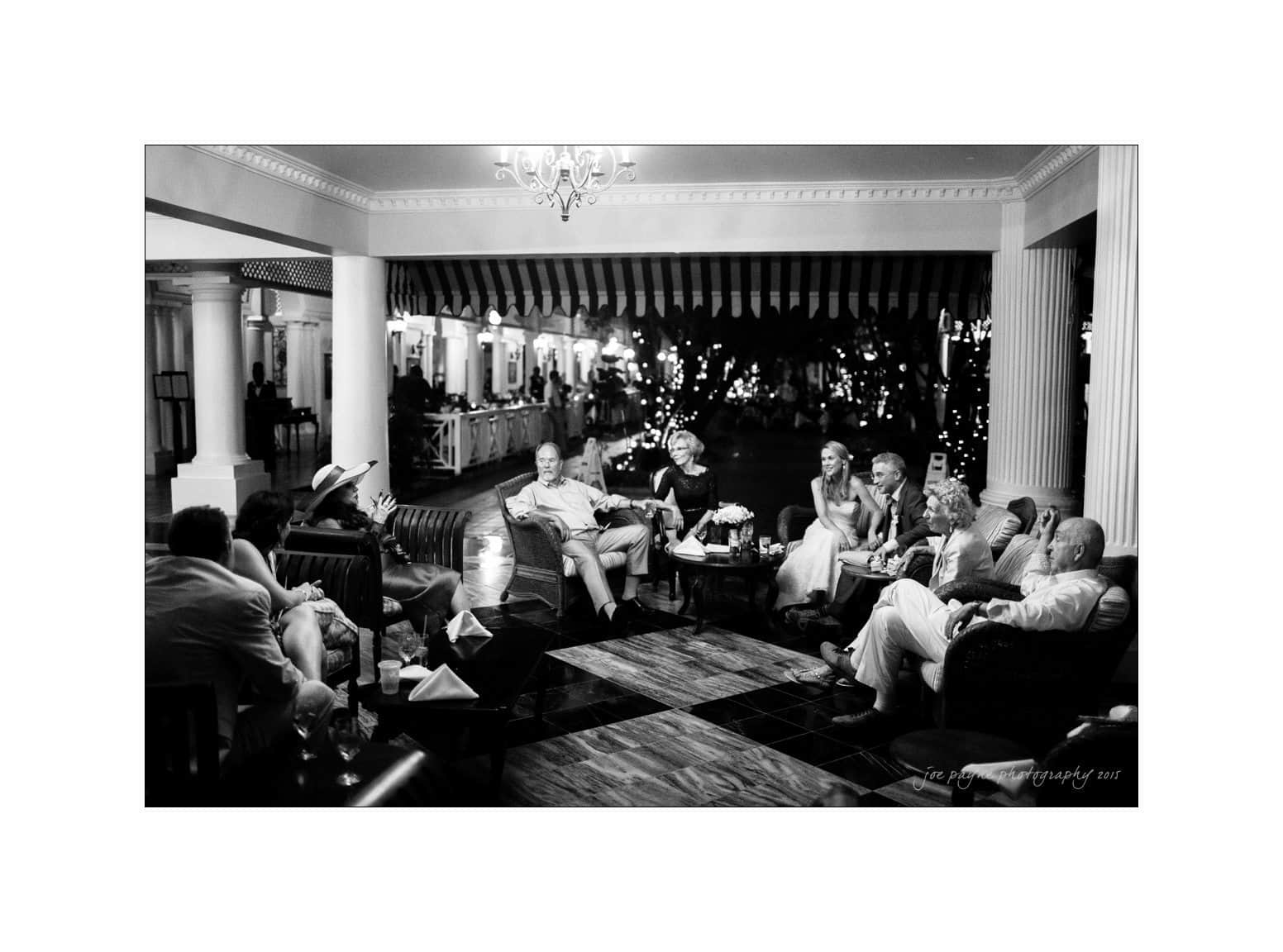 A group of people sit in a well-lit, covered patio area at night. They are engaged in conversation, surrounded by decorative columns and an elegant chandelier above. String lights outside add a festive ambiance.