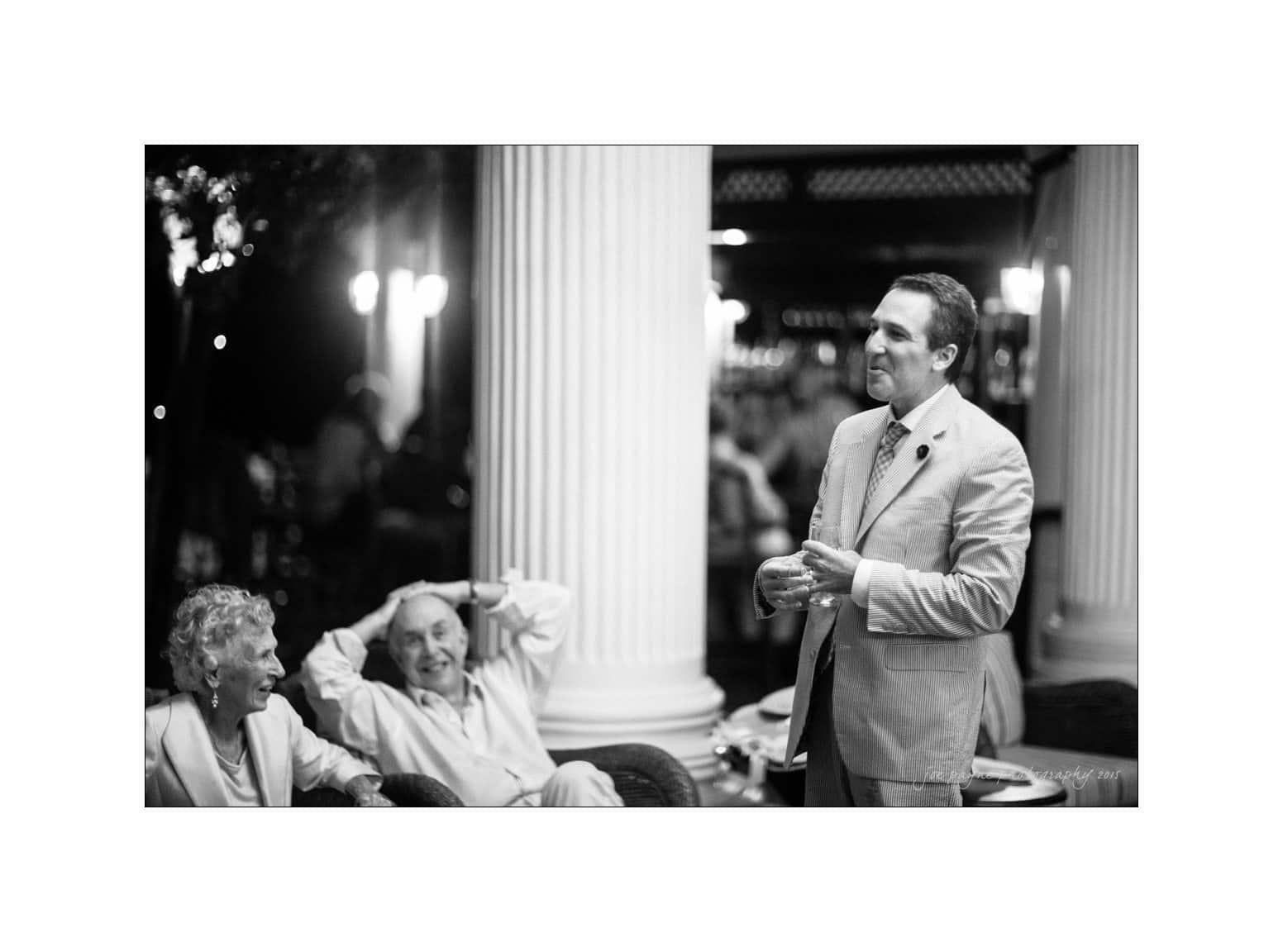 A person in a suit stands speaking, holding a drink, in front of seated elderly individuals. The setting appears to be a formal event with large pillars and ambient lighting. The seated individuals are smiling and relaxed.