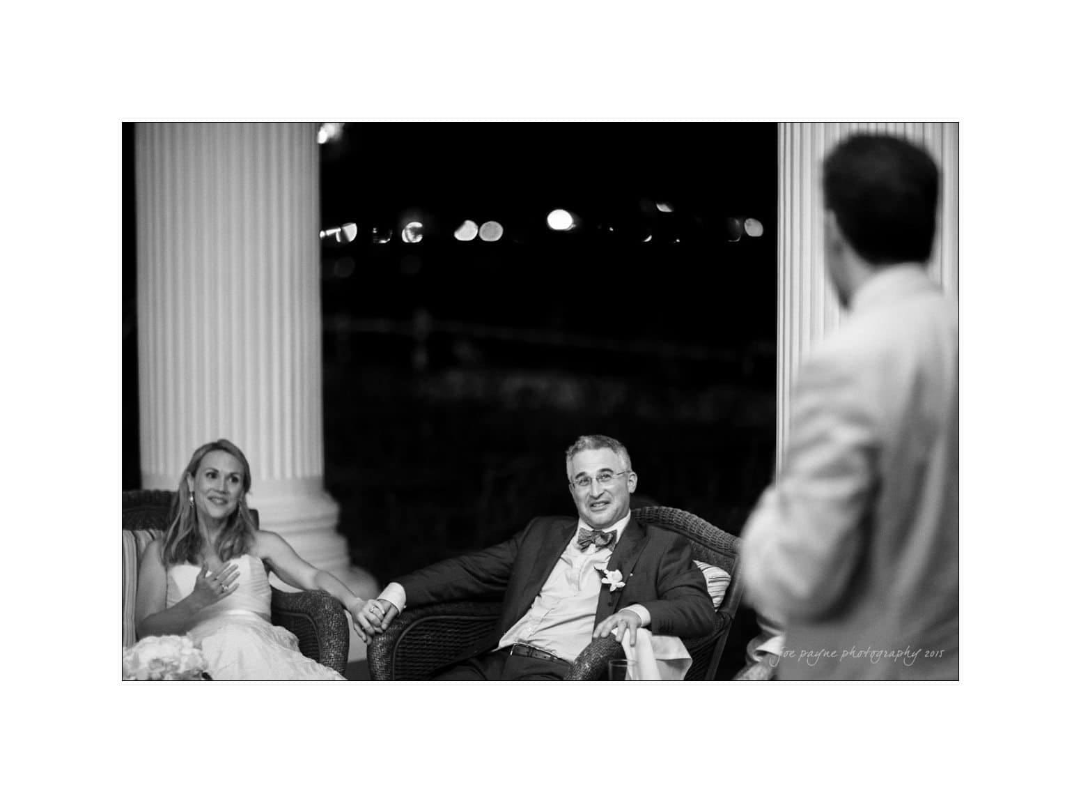 A bride and groom sit holding hands and smiling while listening to a speech. The groom is in a suit, and the bride is in a wedding dress. They are seated on a veranda with columns. A person in a suit stands in the foreground, partially visible.