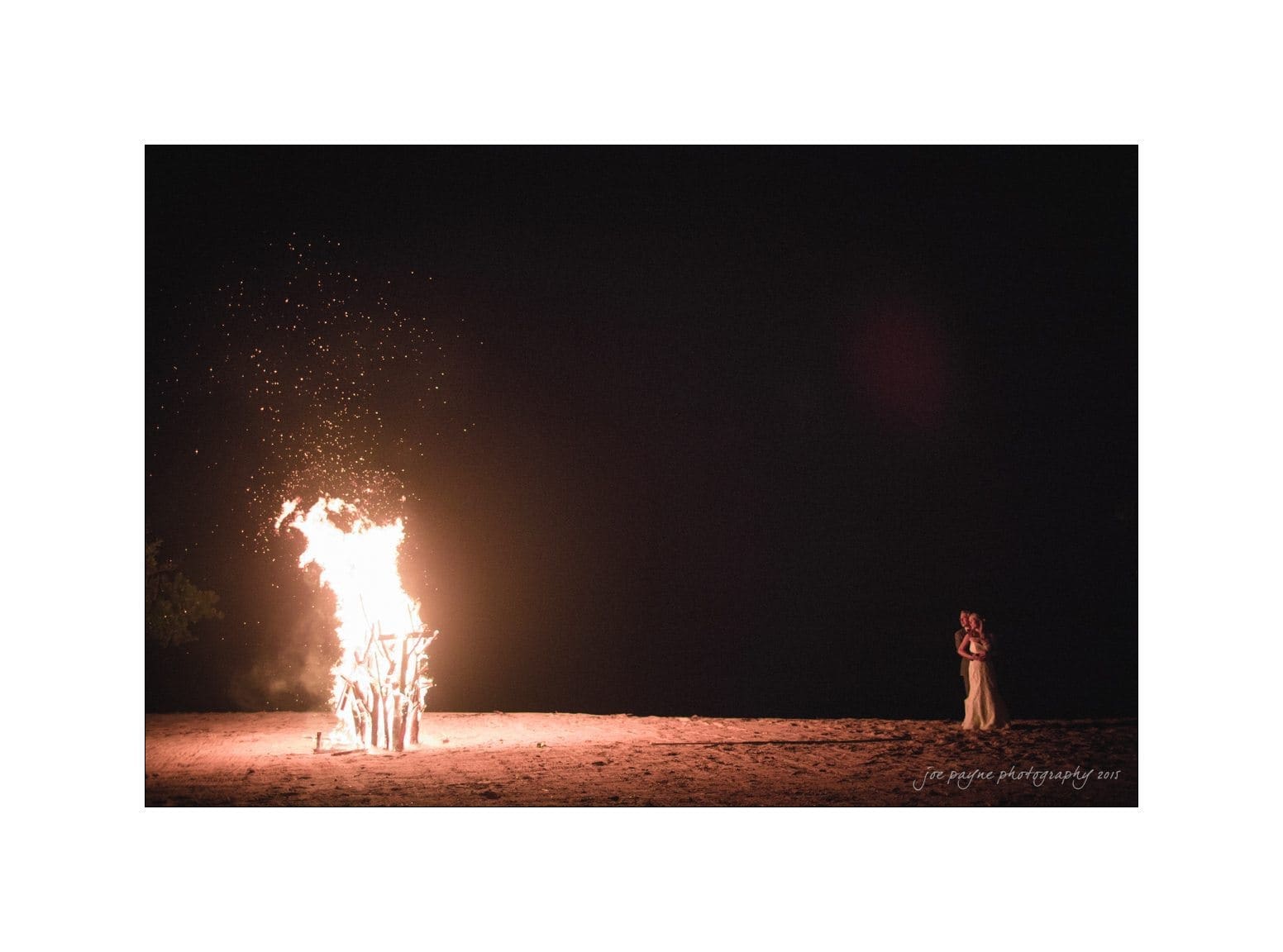 A large bonfire illuminates the night on a beach, with sparks flying upwards. A person stands at a distance, silhouetted against the dark sky, observing the fire.