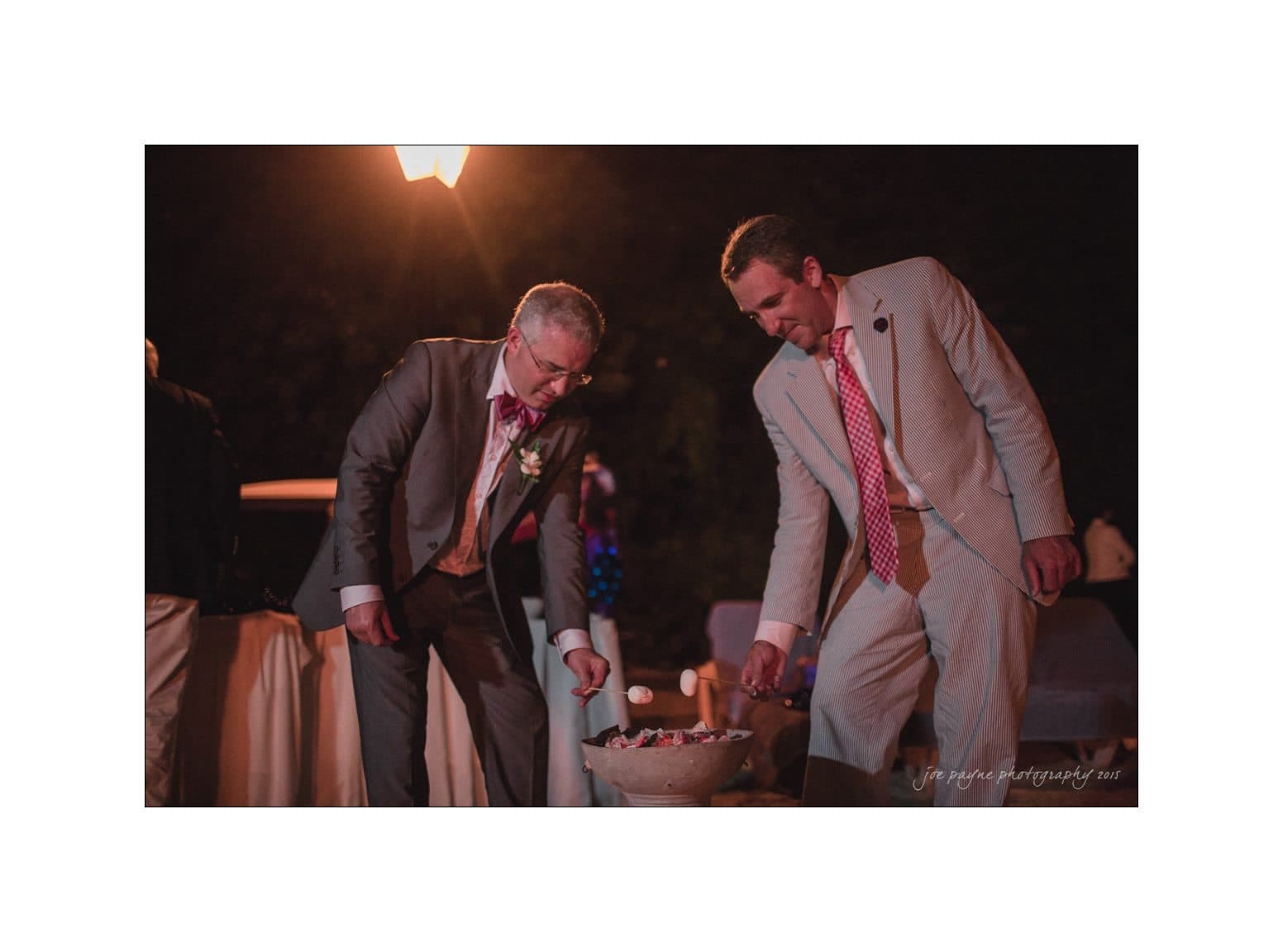 Two men in suits roast marshmallows over a small fire pit at an outdoor evening event. The setting is warmly lit, creating a cozy atmosphere.