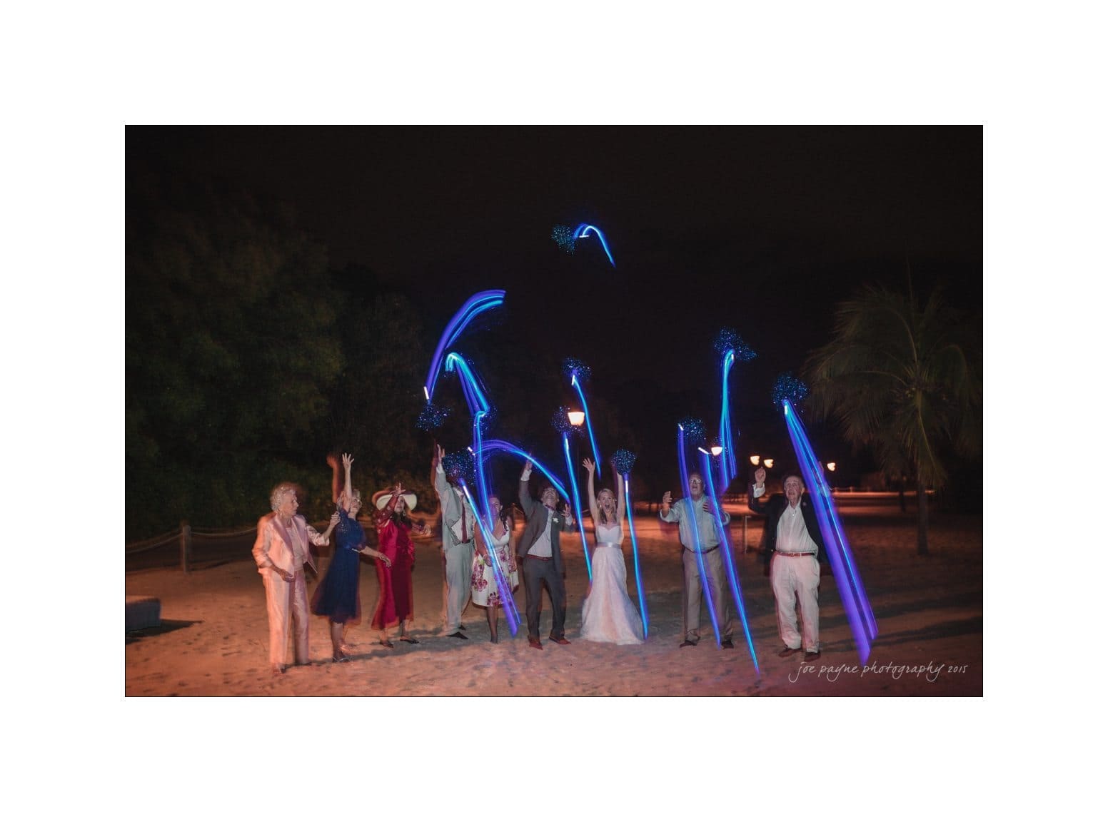 A group of people stands on a sandy area at night, cheerfully waving blue light sticks in the air. Trees and dim lights are visible in the background. They appear to be celebrating an event.