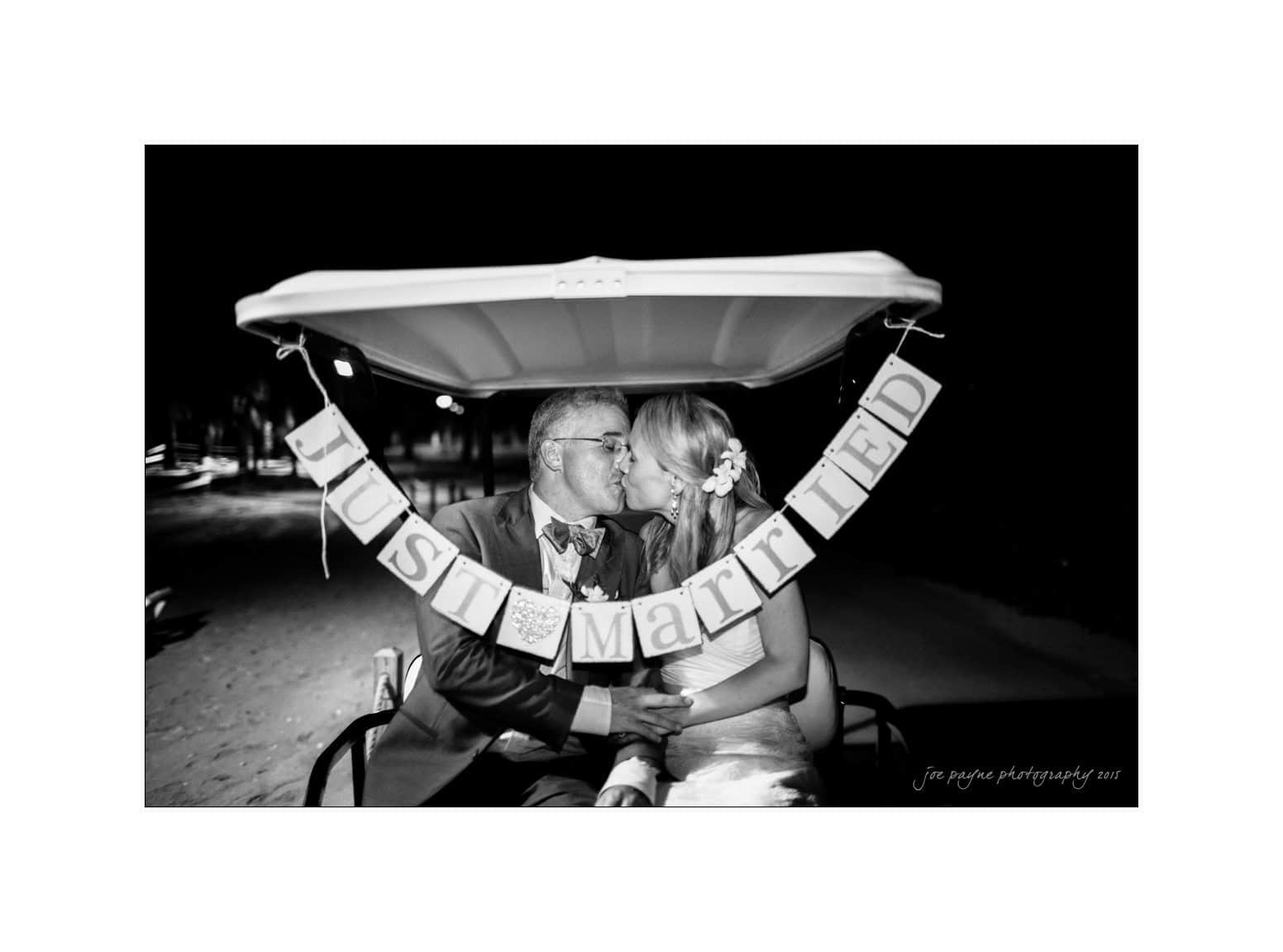 half moon montego bay wedding photography - leah & evan A newlywed couple sits on a golf cart, kissing. A Just Married banner hangs on the carts back. The photo is in black and white, capturing a nighttime scene.