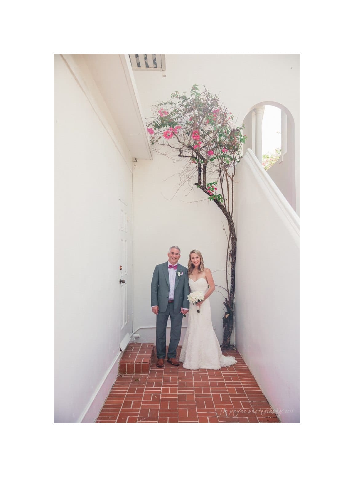 A bride in a white gown and groom in a gray suit stand under a flowering tree against a white wall. They are smiling and holding hands on a brick path, with a staircase and windows visible on the right.
