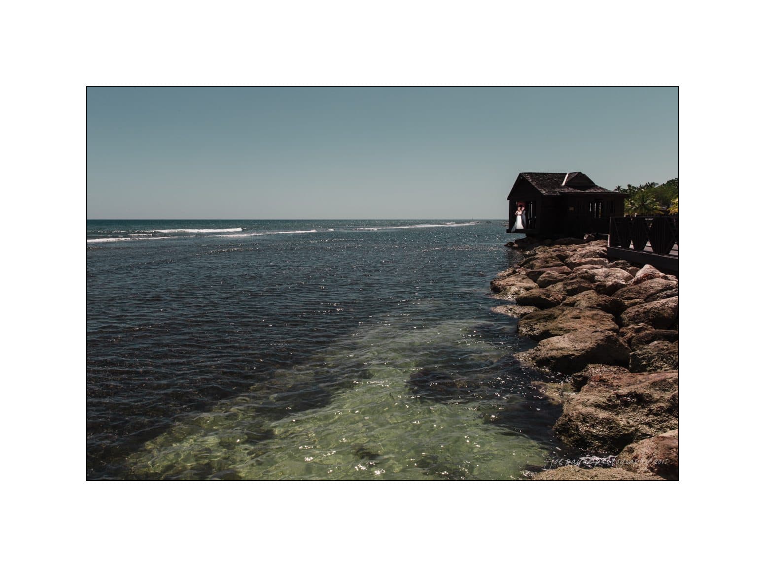 half moon montego bay wedding photography - leah & evan A wooden gazebo perched on a rocky shoreline overlooks a vast, calm ocean under a clear blue sky. Sunlight creates a greenish hue in the shallow water near the rocks.