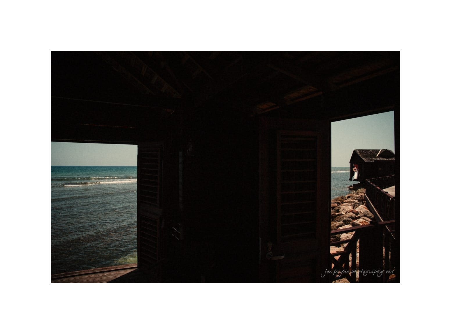 A dimly lit room with open wooden shutters looks out onto a view of the ocean. Sunlight streams in, illuminating part of the interior. The horizon and gentle waves are visible through the open window.