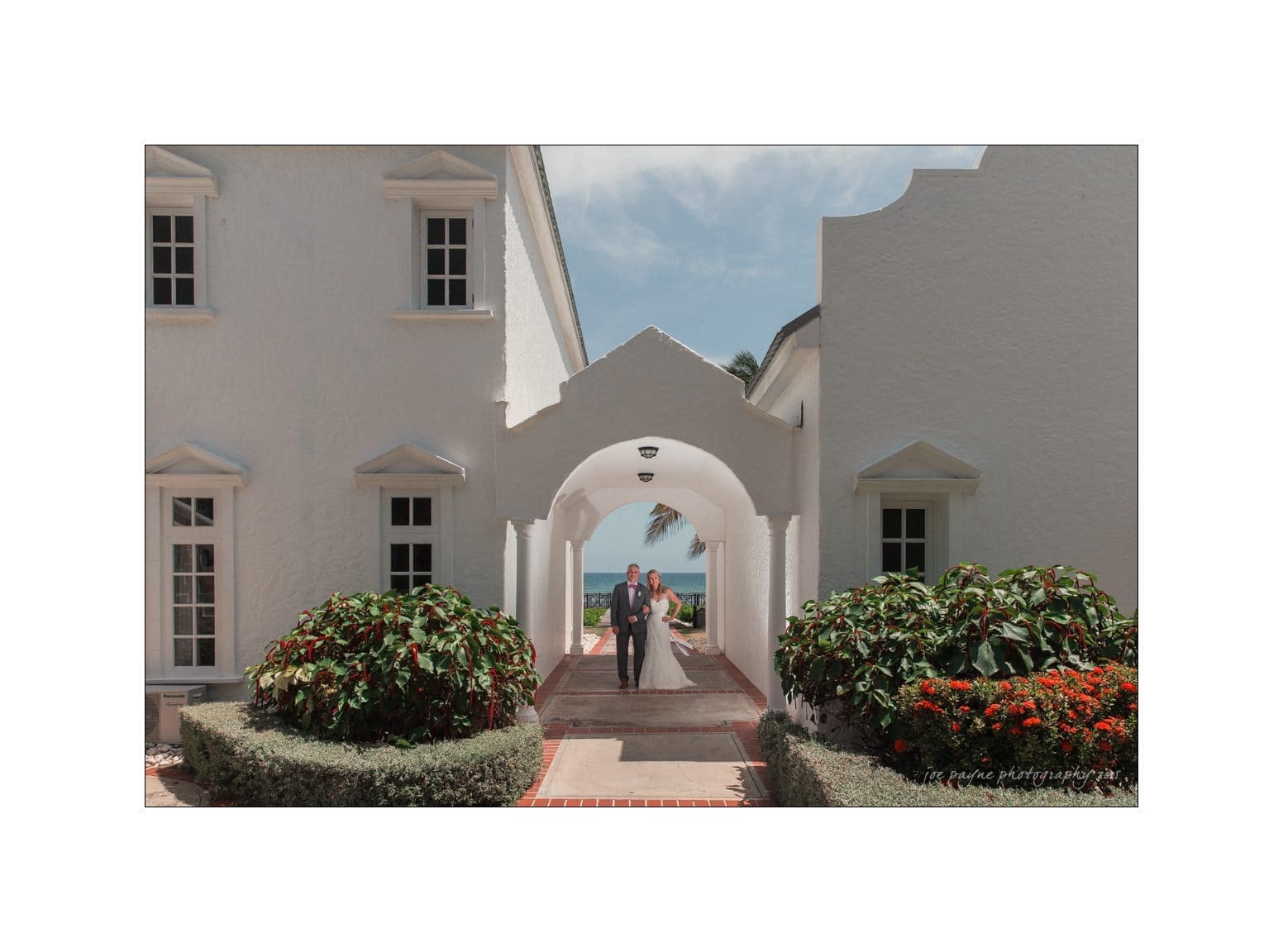 A couple stands together under an archway in a white building with a view of the ocean in the background. Lush green plants and red flowers border the walkway, creating a picturesque, tropical setting.