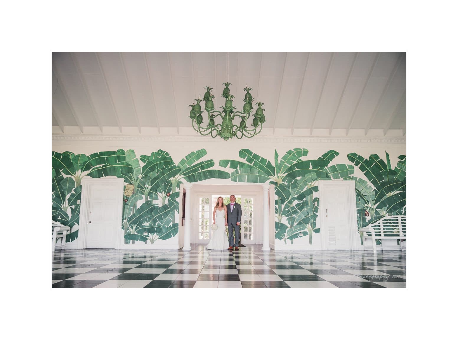 A bride and groom stand in an elegant room with a bold tropical leaf mural and a large green chandelier. The floor features a black and white checkered pattern, and doors with glass panels flank the couple.