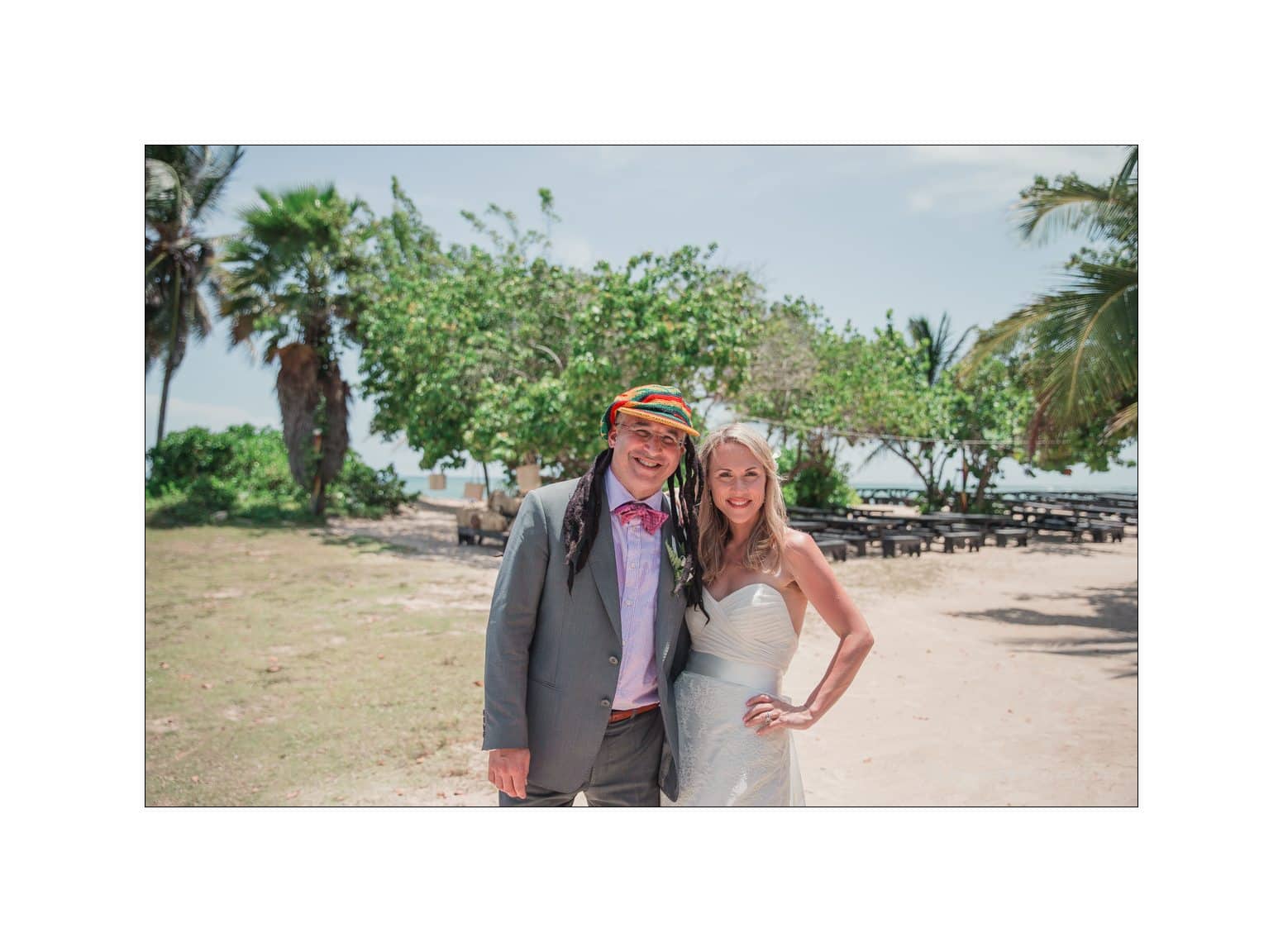 A smiling couple stands together outdoors on a sunny day, surrounded by greenery. The man wears a colorful hat and a suit, while the woman wears a strapless white dress. Palm trees and picnic tables are visible in the background.
