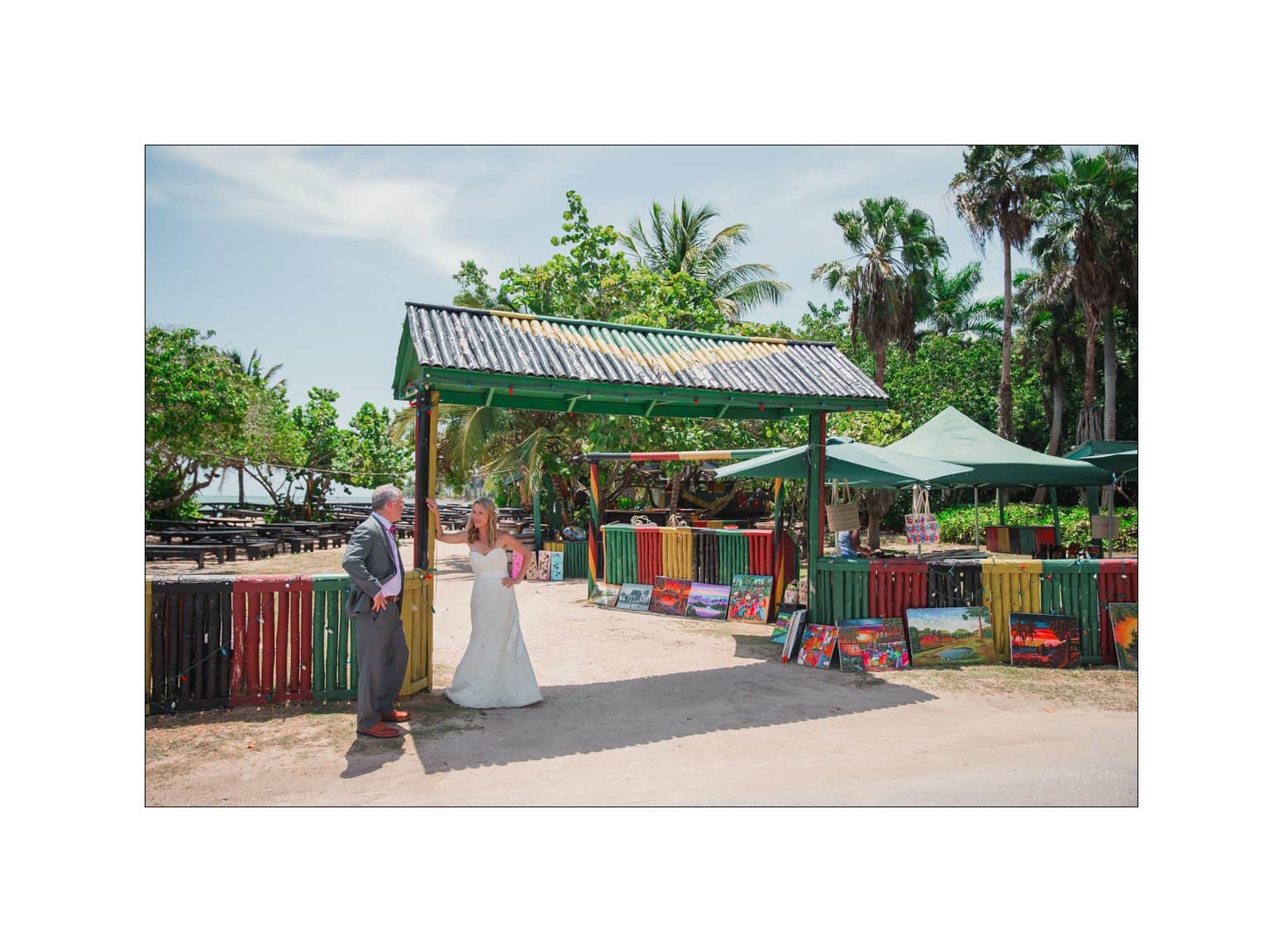 A bride and groom stand in front of a colorful, open-air art market. The groom, in a gray suit, and the bride, in a white gown, are surrounded by colorful paintings. The market has a rustic shelter and vibrant fenced display, with tropical trees nearby.