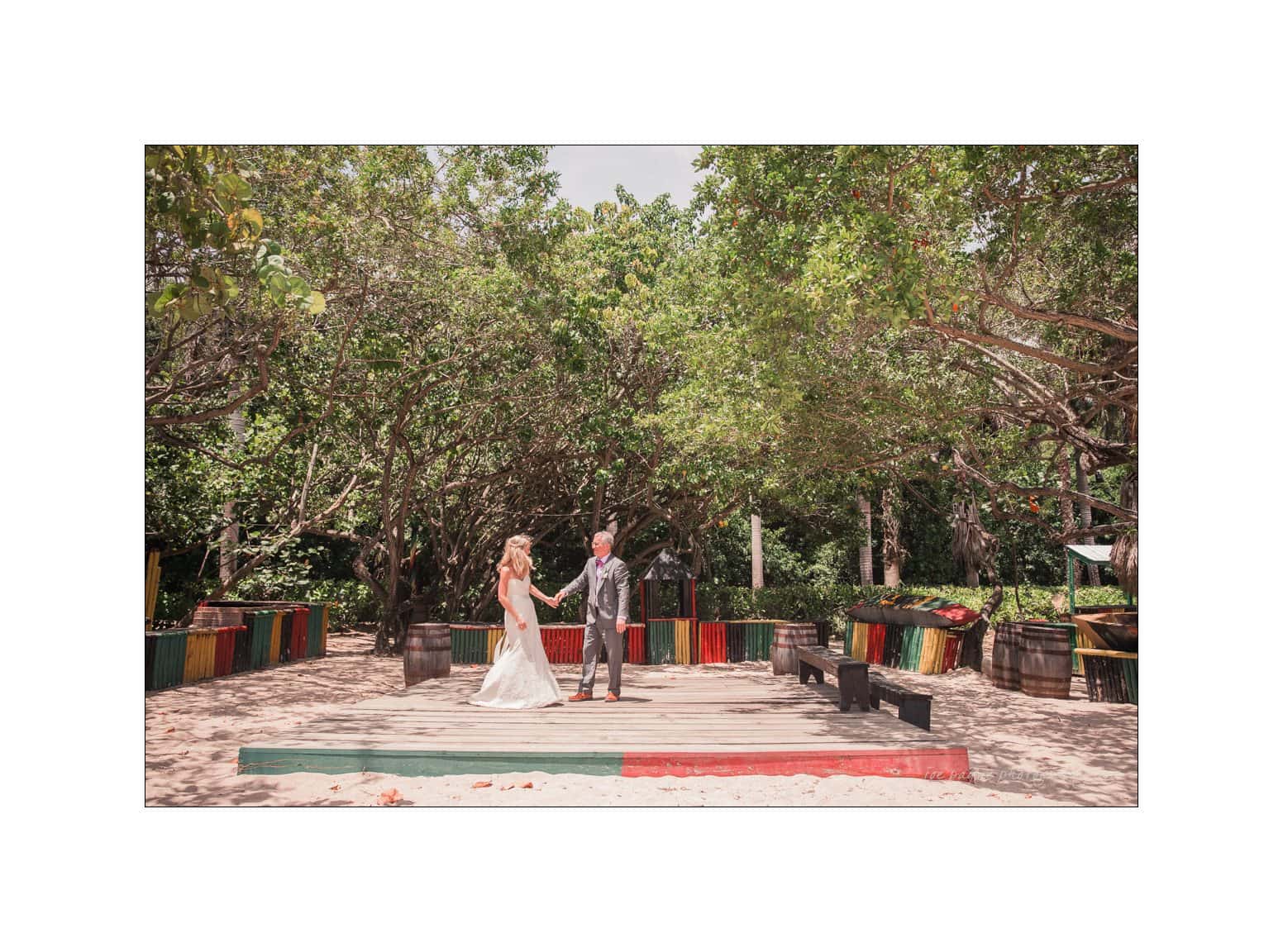 half moon montego bay wedding photography - leah & evan A couple stands on a wooden platform in a lush outdoor setting. They hold hands, surrounded by trees and colorful wooden railings. The bride wears a white gown, and the groom is in a gray suit. Sunlight filters through the tree canopy.