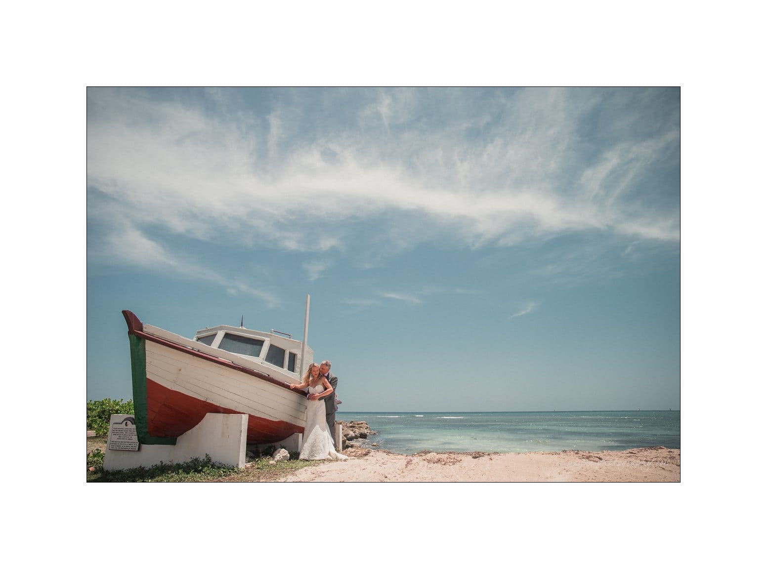 A couple embracing near a beached wooden boat on a sandy shore, under a blue sky with wispy clouds. The ocean is calm in the background.