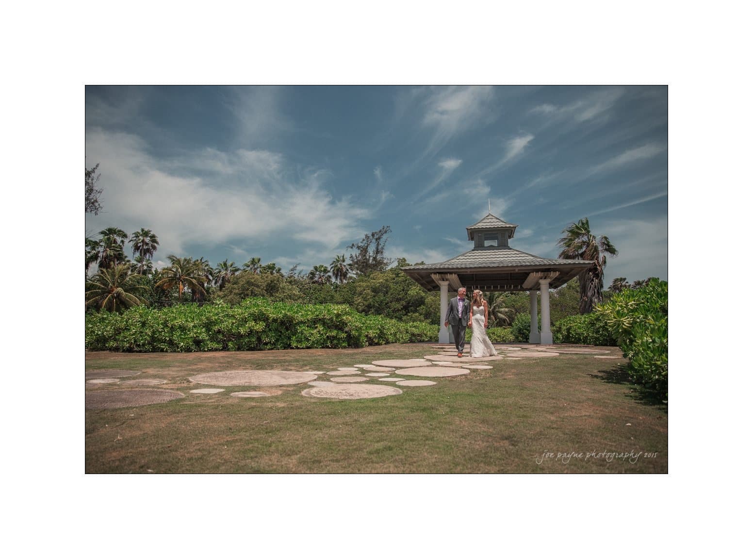 A couple walks hand in hand down a stone path toward a gazebo surrounded by lush greenery and palm trees under a partly cloudy sky.