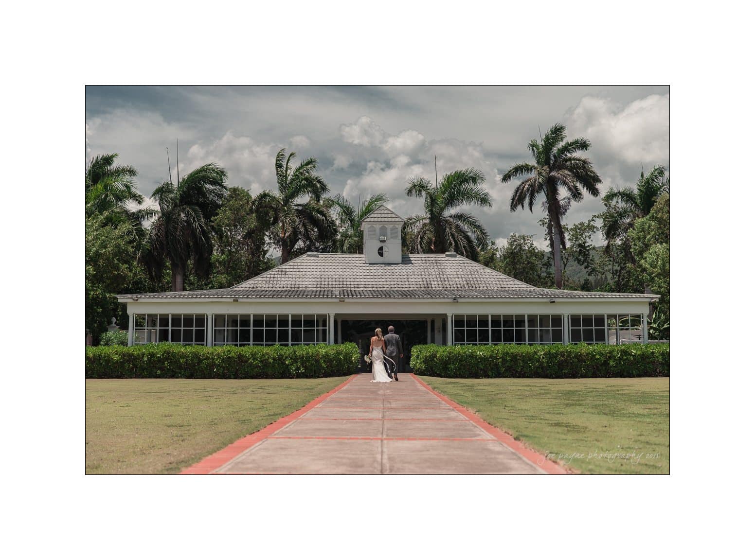 A bride and groom walk hand-in-hand towards a white church with a clock tower, surrounded by palm trees under a cloudy sky. The path is lined with grass, creating a serene and picturesque setting.