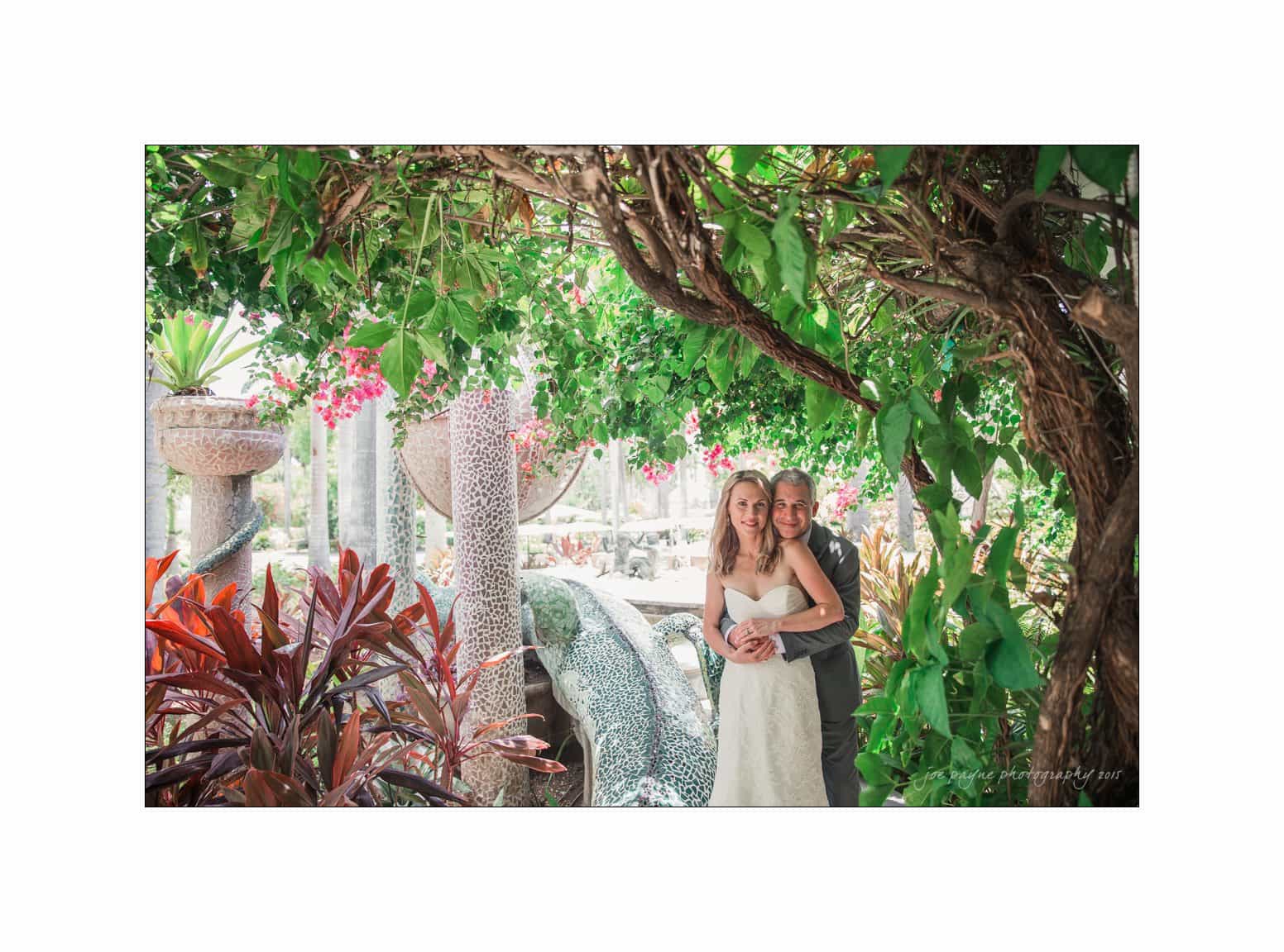 half moon montego bay wedding photography - leah & evan A couple embraces in a lush, colorful garden. The woman is wearing a white dress, and the man is in a suit. They are surrounded by vibrant plants and mosaic sculptures, with sunlight filtering through the foliage above.