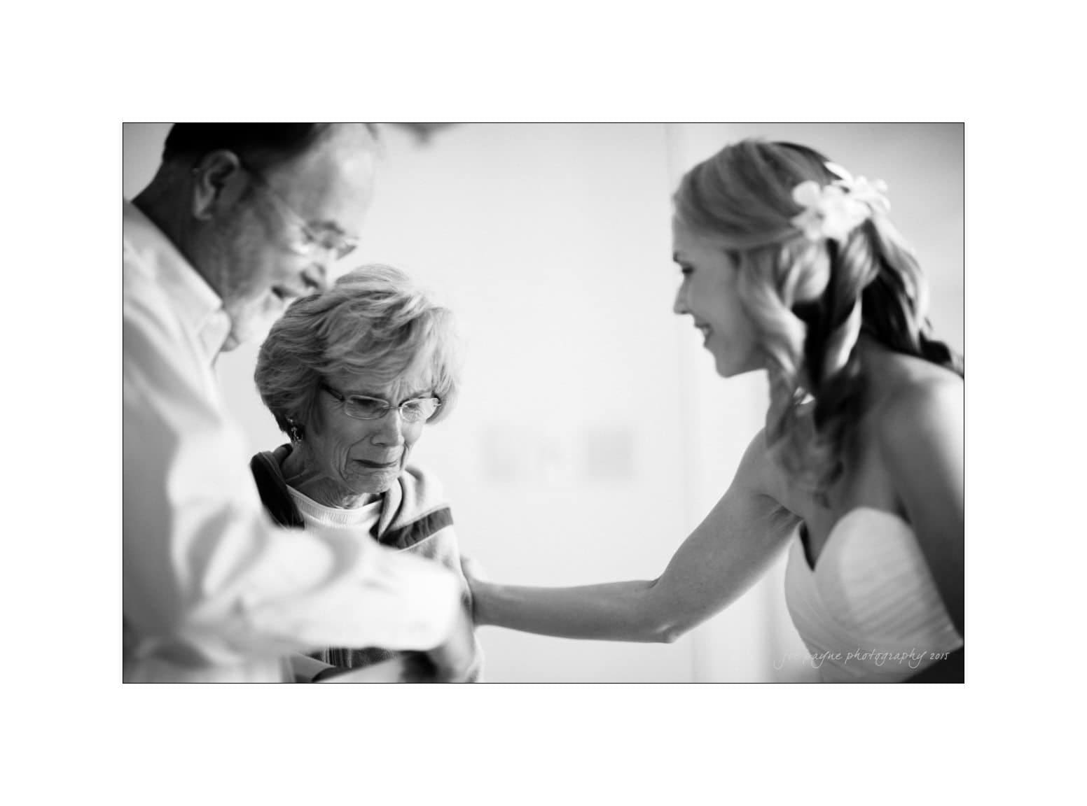A black and white image of three people in a joyful moment. A woman in a wedding dress smiles, reaching out towards an elderly woman wearing glasses, who is standing next to a man with a beard.
