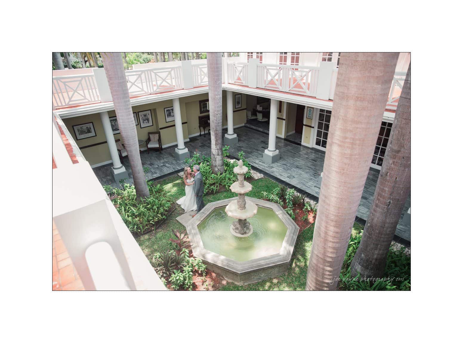 A person stands near a central stone fountain in a lush garden courtyard surrounded by tall palm trees and a two-story building with balconies. The atmosphere is peaceful and picturesque.