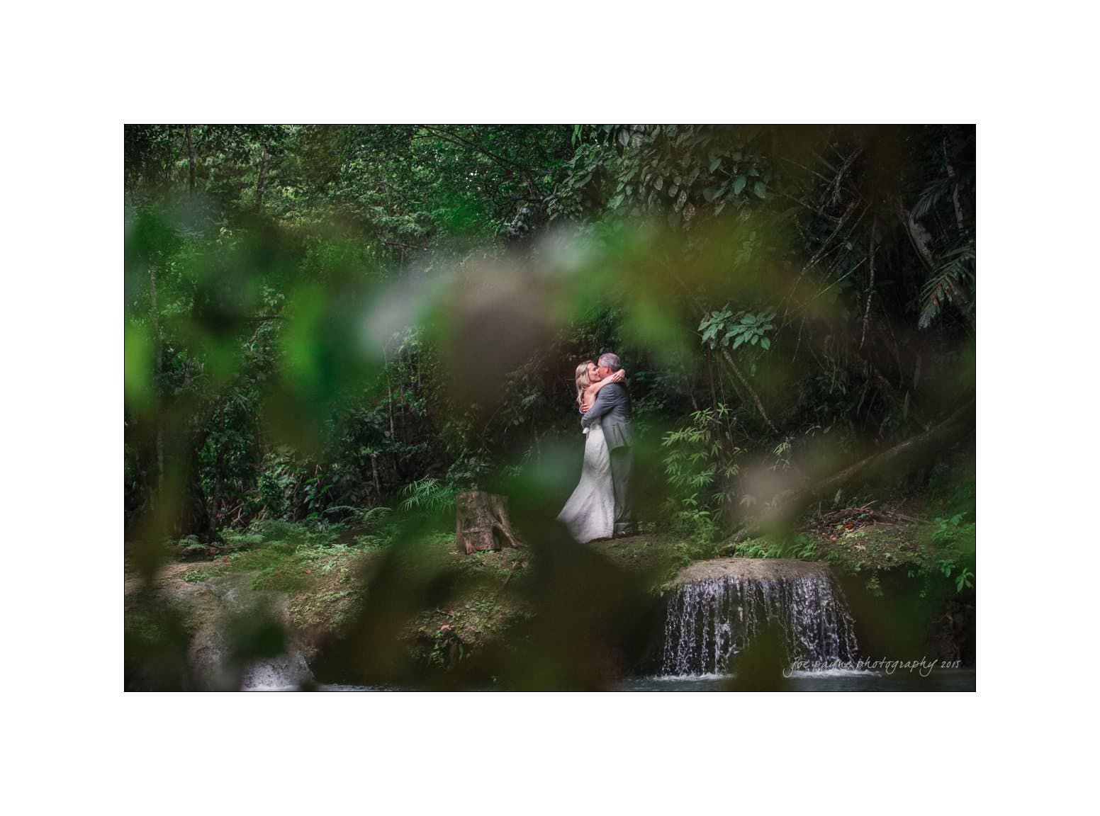 A couple embraces in a lush forest setting. They stand near a small waterfall surrounded by greenery, creating a serene and romantic atmosphere. The view is partially obscured by leaves, adding a sense of depth and intimacy to the scene.