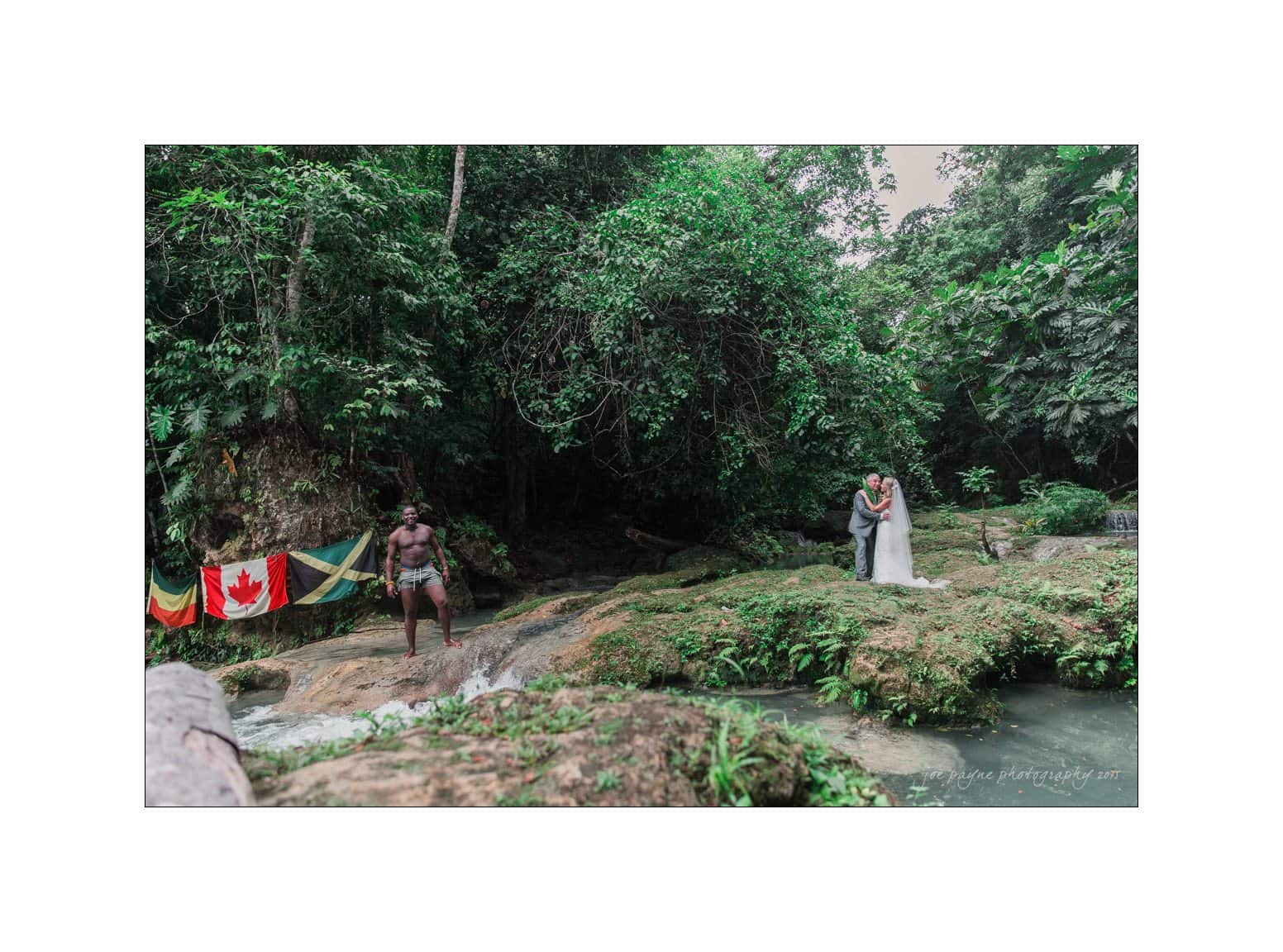 half moon montego bay wedding photography - leah & evan A wedding couple stands on a lush, green rock beside a flowing stream in a tropical forest. Flags from Canada and Ethiopia are hung nearby. A man dressed in traditional attire stands on another rock.