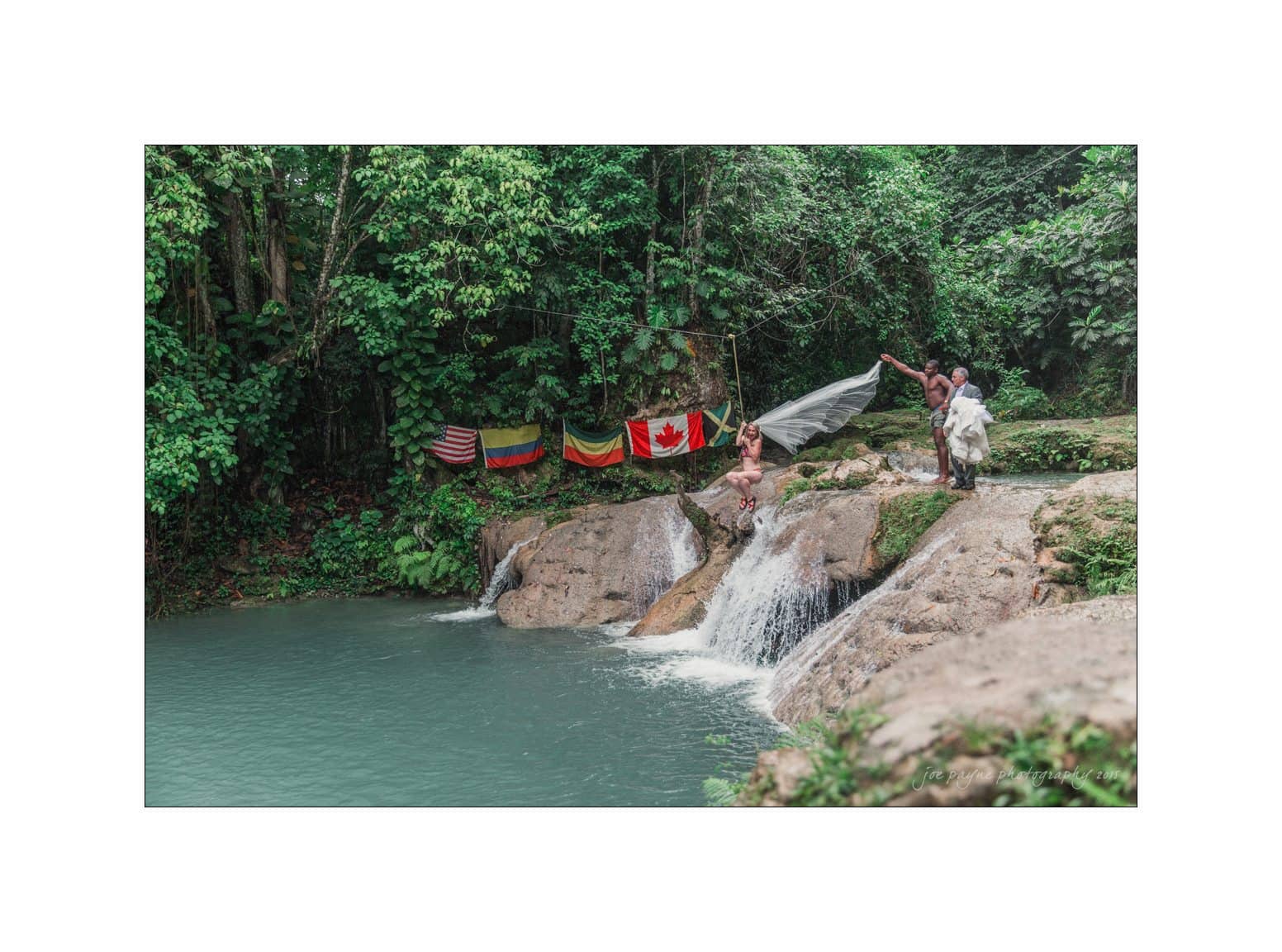 Two people enjoy a waterfall with lush greenery around them. Flags of the USA, Colombia, and Canada are hung in the background, adding colorful accents to the natural scene.