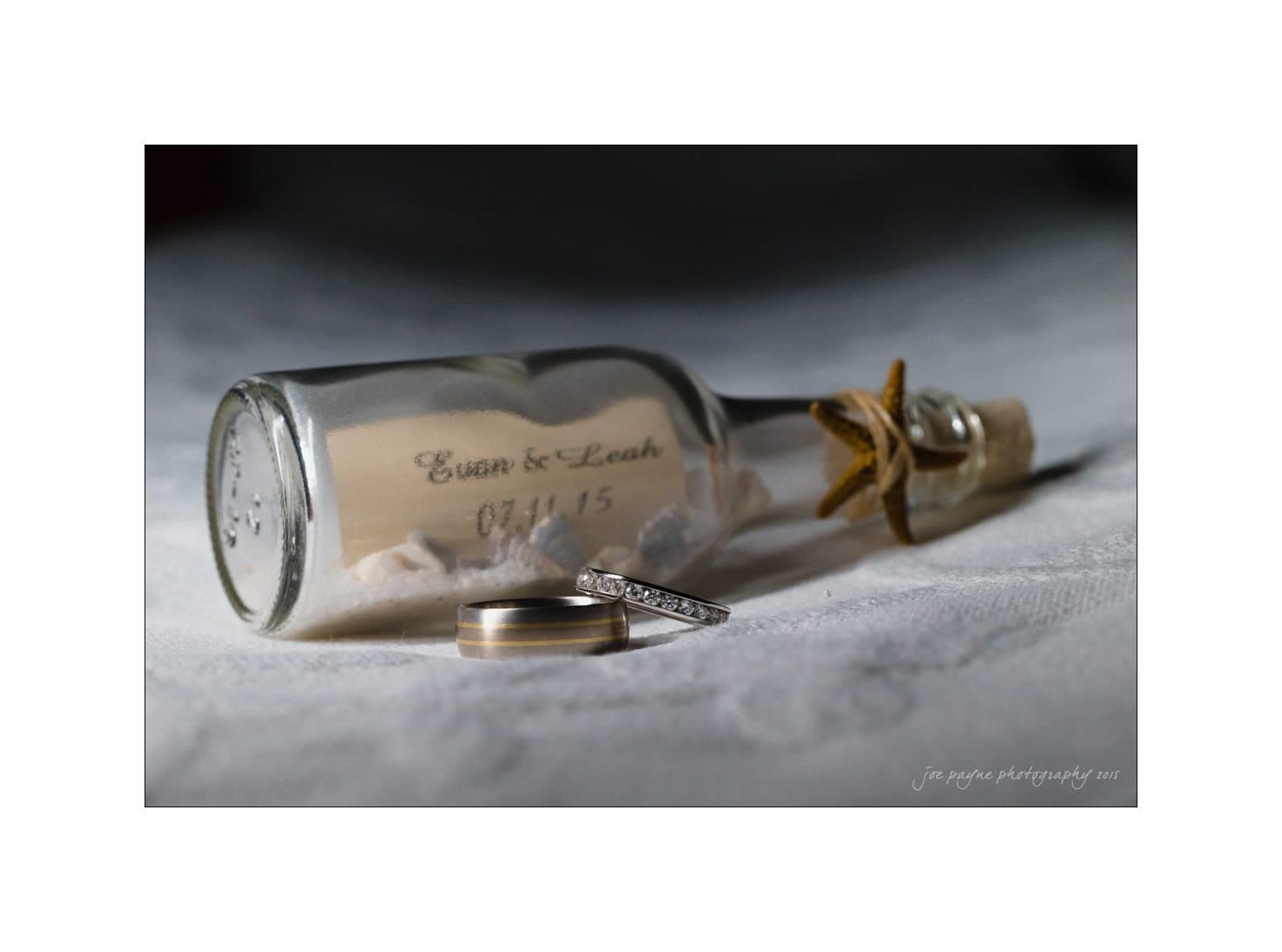 A decorative bottle lies on sand with embossed text Evan & Leah 07.11.15. Two rings rest nearby, one with a diamond. A starfish is attached to the bottles neck.