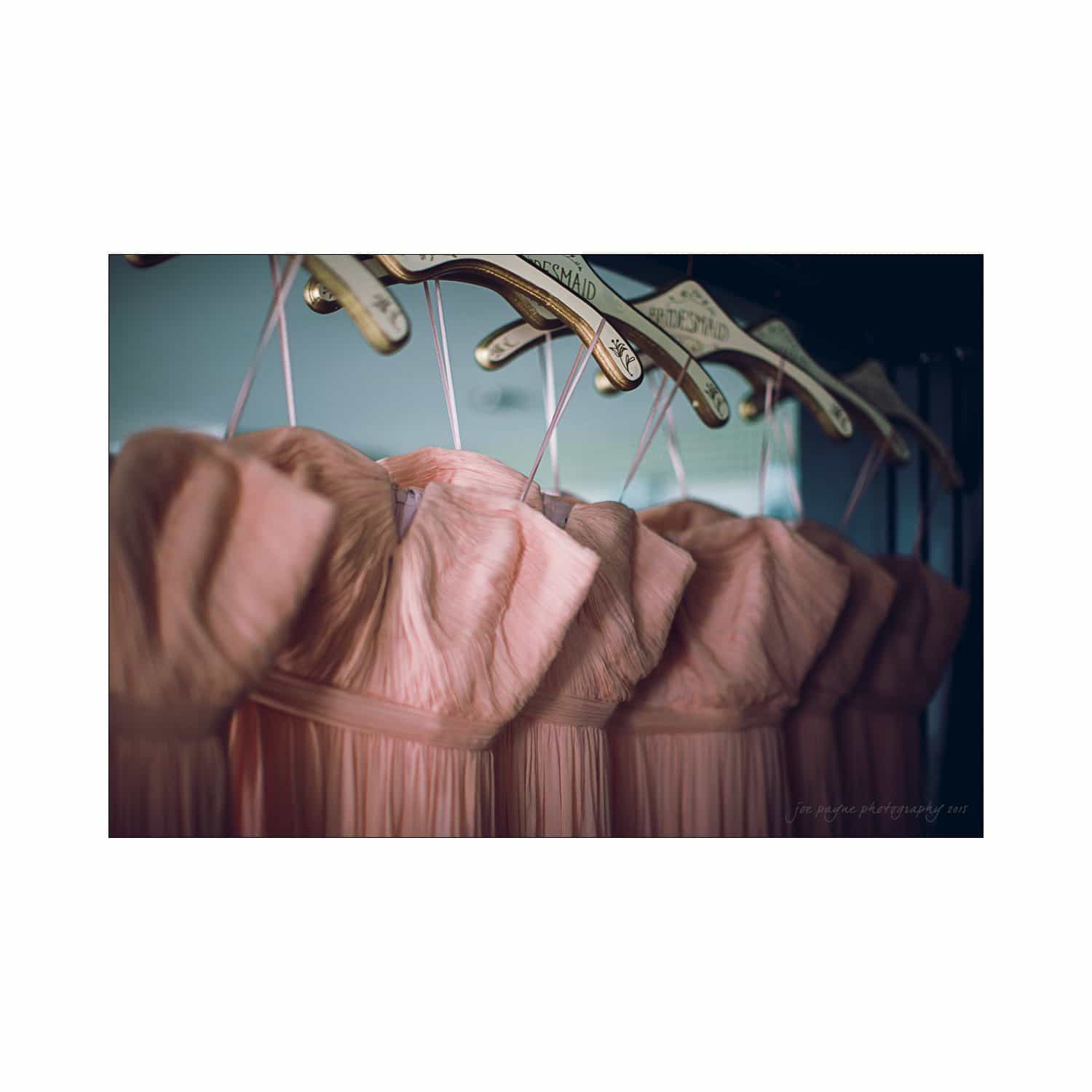 A row of peach-colored, strapless bridesmaid dresses hanging on personalized wooden hangers. The soft fabric and gentle pleats create a delicate and elegant appearance.