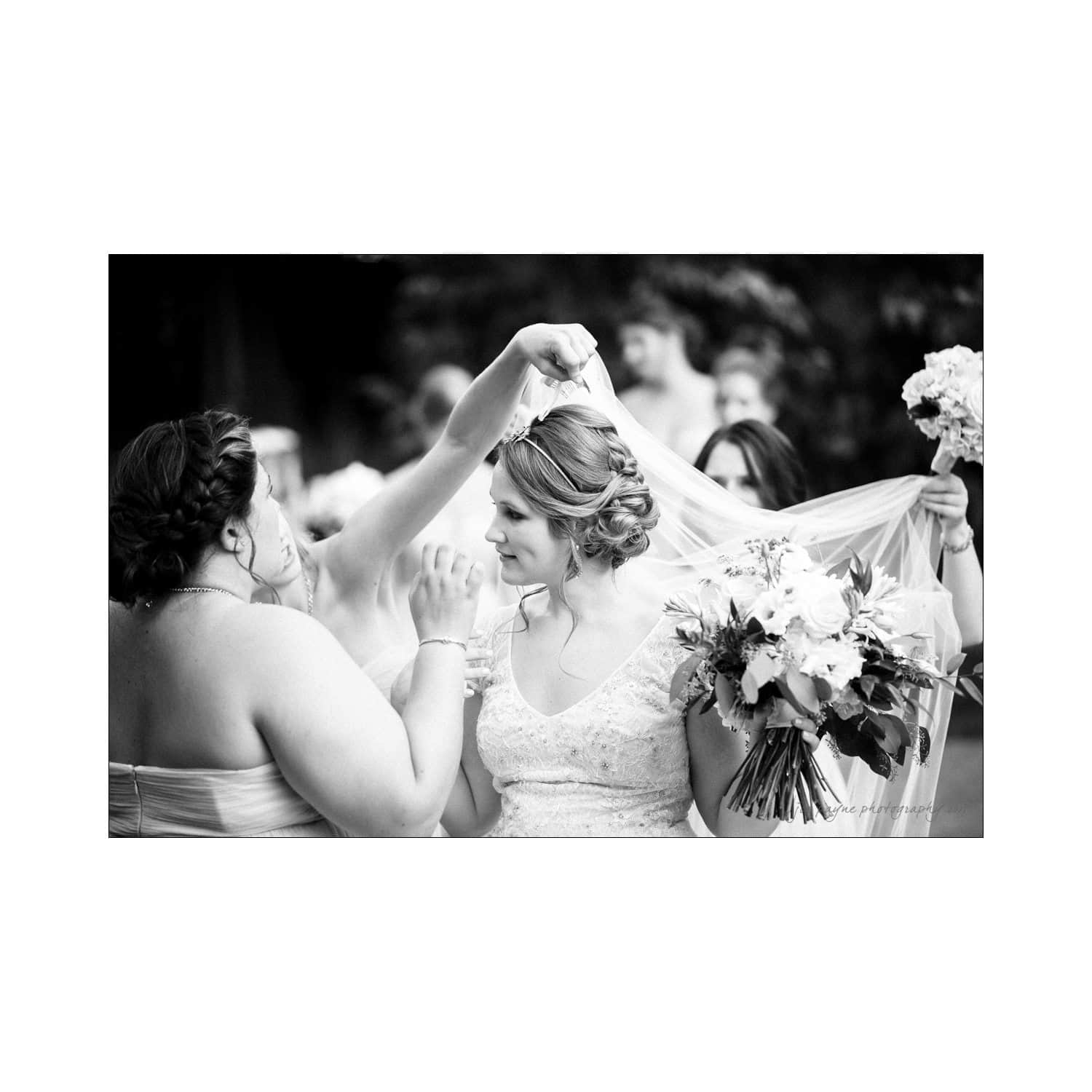 A black and white photo of a bride holding a bouquet, while another woman adjusts her veil. The scene is joyful, surrounded by bridesmaids and greenery in the background.