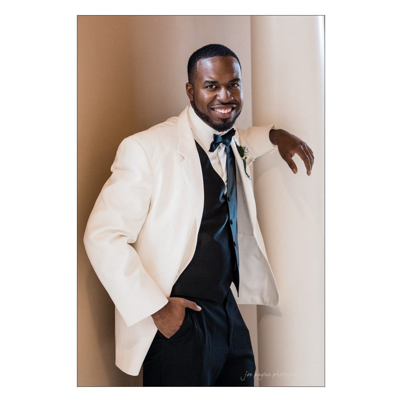 A man in a white suit jacket and dark vest smiles while leaning against a wall. His left hand is in his pants pocket, and he wears a bow tie with a small boutonniere pinned to his jacket.