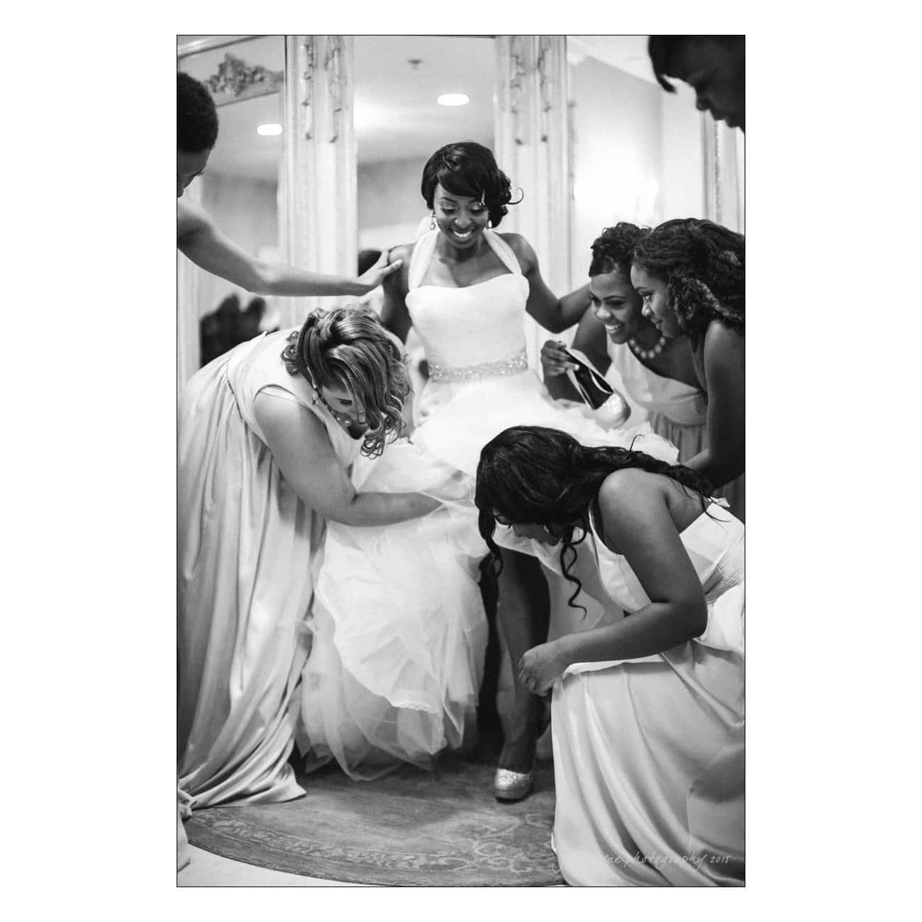 A bride in a wedding dress joyfully sits while surrounded by her bridesmaids. They help adjust her outfit and shoes. The scene is filled with happiness and camaraderie as they prepare for the ceremony in a warmly lit room.