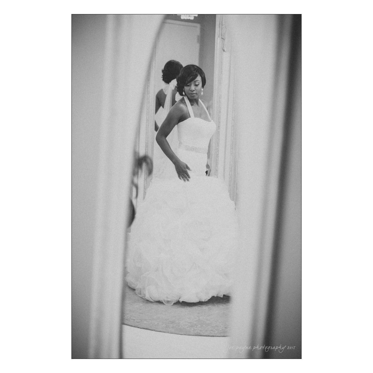 A bride in a wedding gown with a halter neckline stands in front of a mirror, adjusting her dress. The image is in black and white, capturing her reflection and expression in a candid moment.